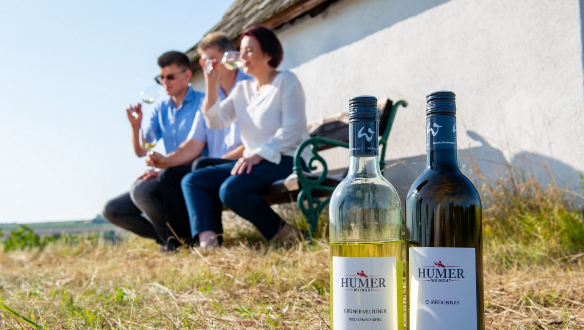 Az előtérben két palack Weingut Humer bor, a háttérben három ember egy padon borozgat.