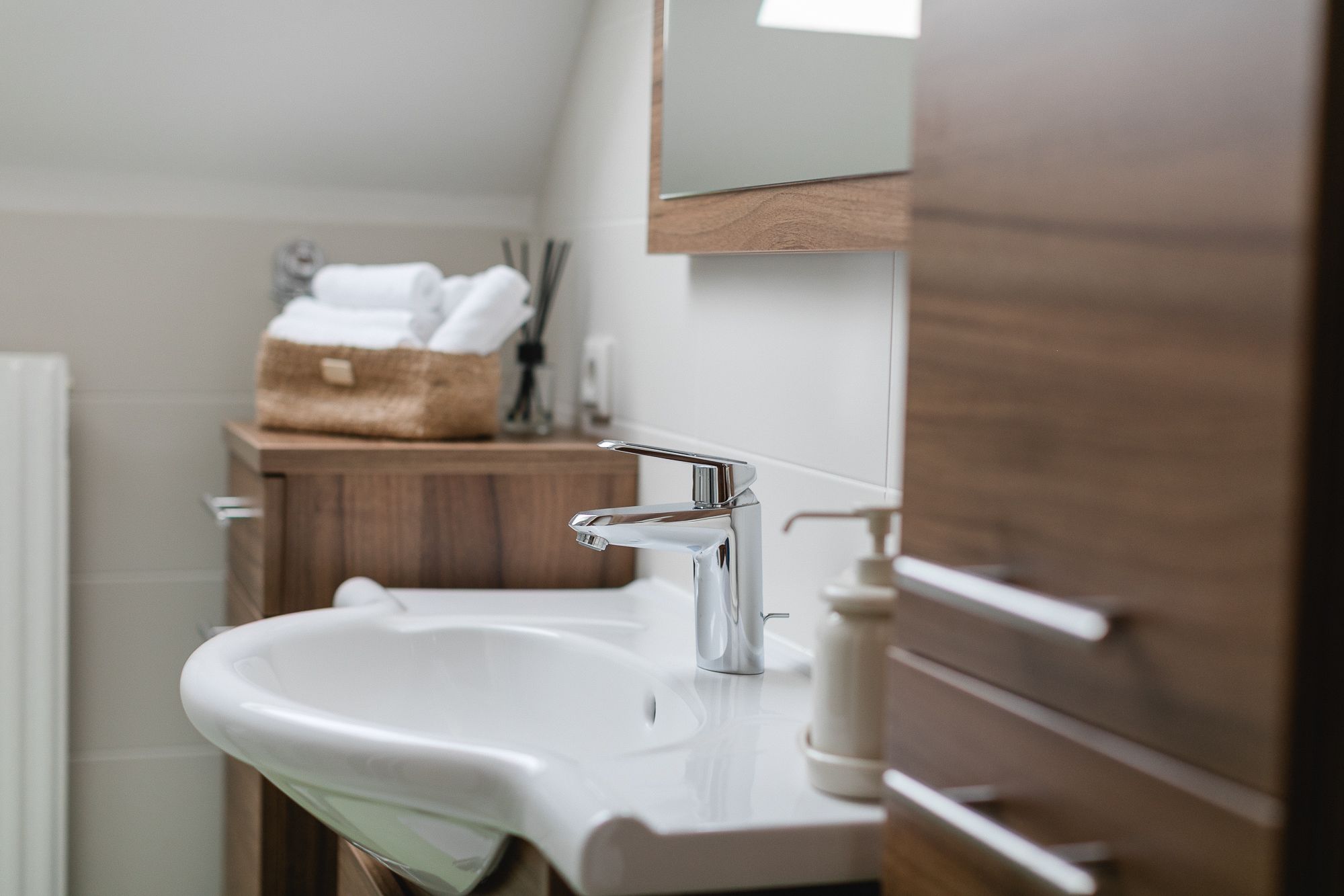 Modern fürdőszoba mosdókagylóval, fából készült szekrényekkel és kosárban elhelyezett törölközőkkel.