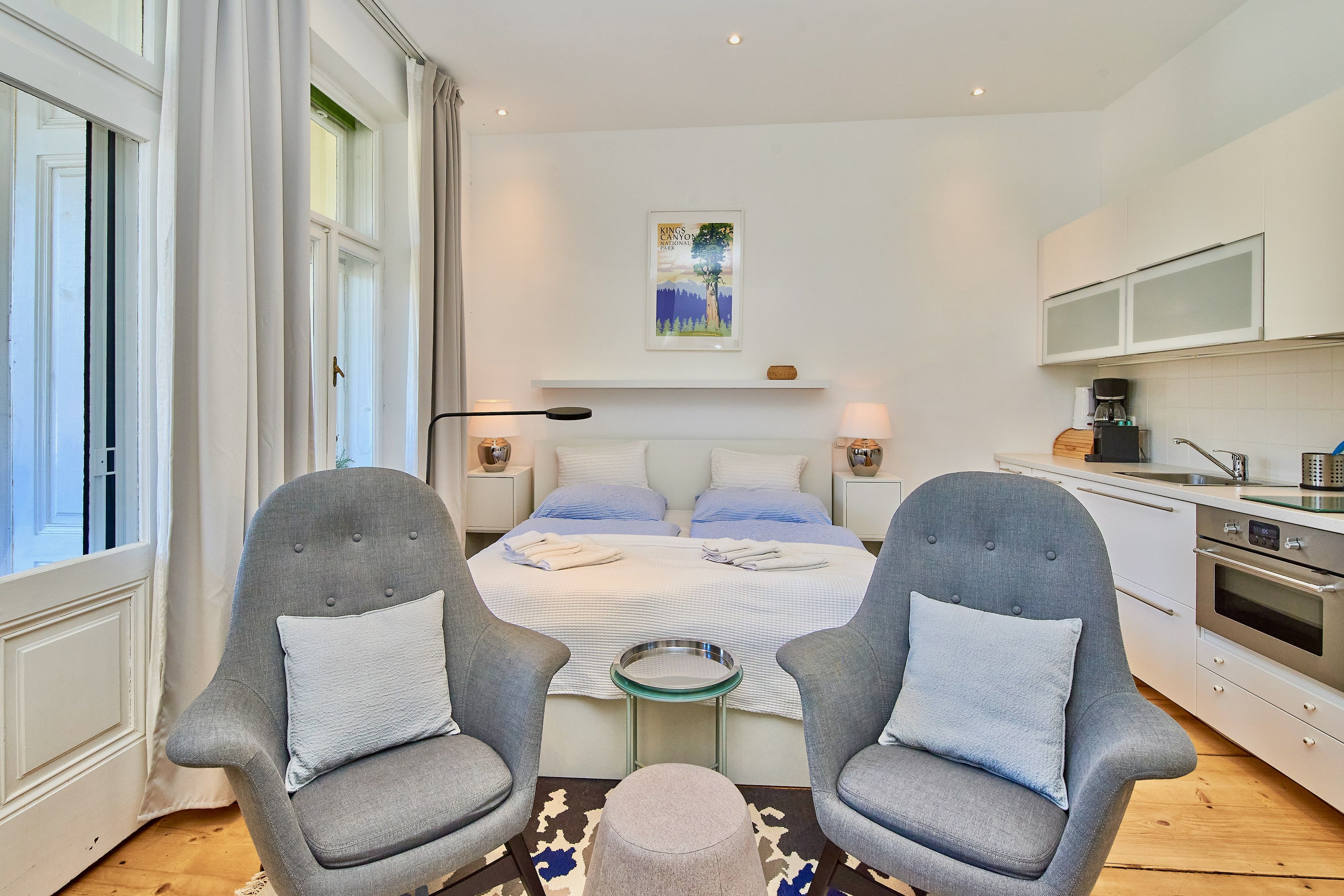 Modern stúdió apartman franciaággyal, két szürke fotel, konyhasarok és nagy ablakok.