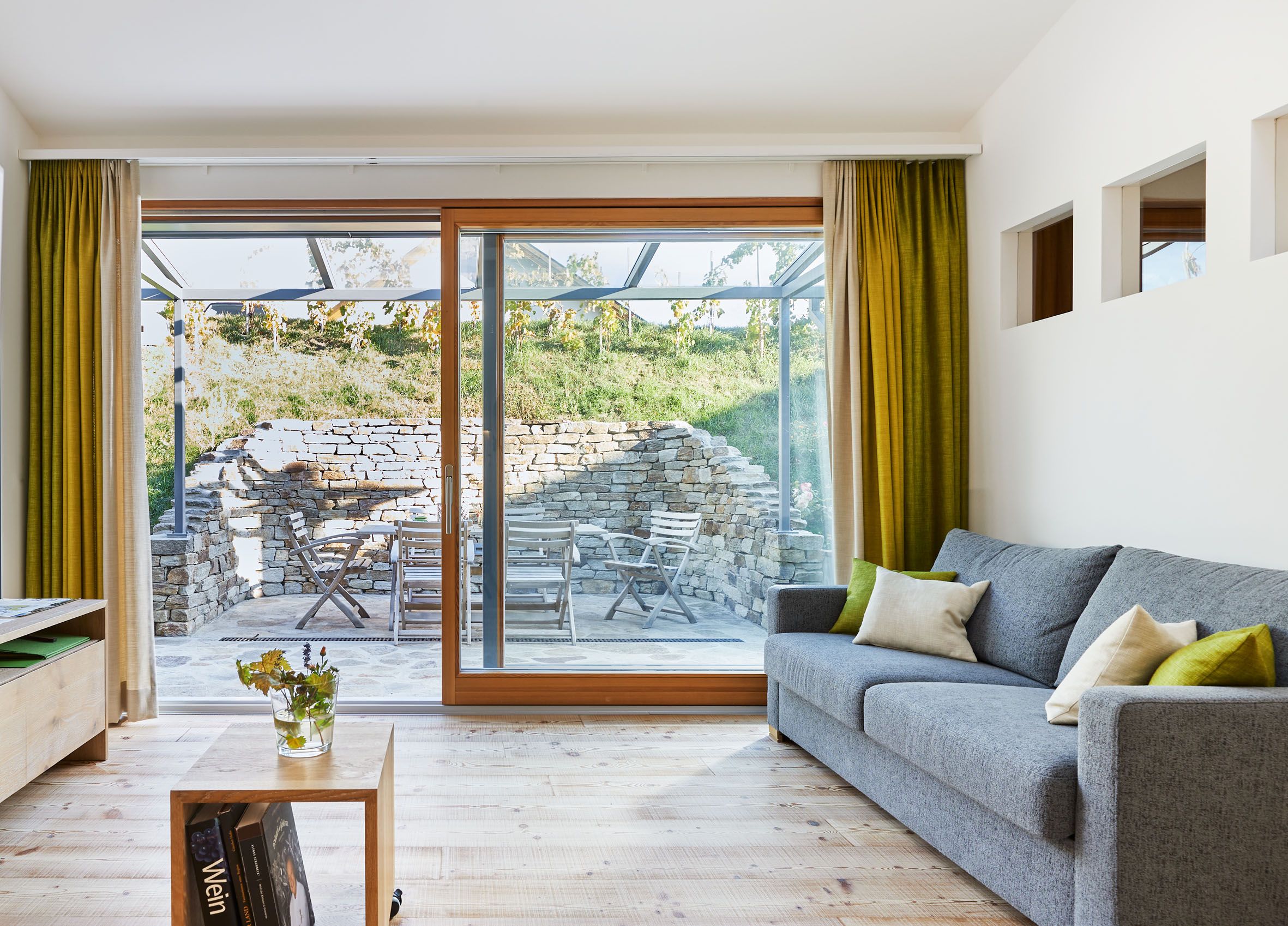 Modern nappali szürke kanapéval, fapadlóval és nagy ablakokkal, amelyek a kőfallal és kerti bútorokkal berendezett teraszra néznek.