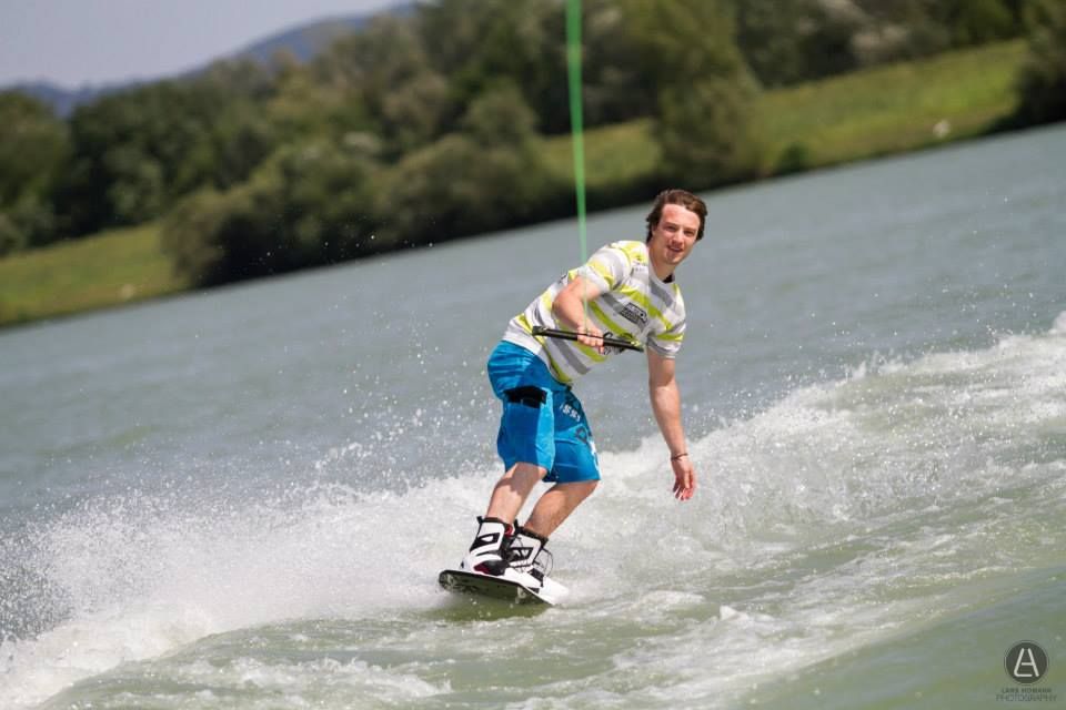 Egy wakeboardos egy zöld tájjal körülvett tavon száguldozik.