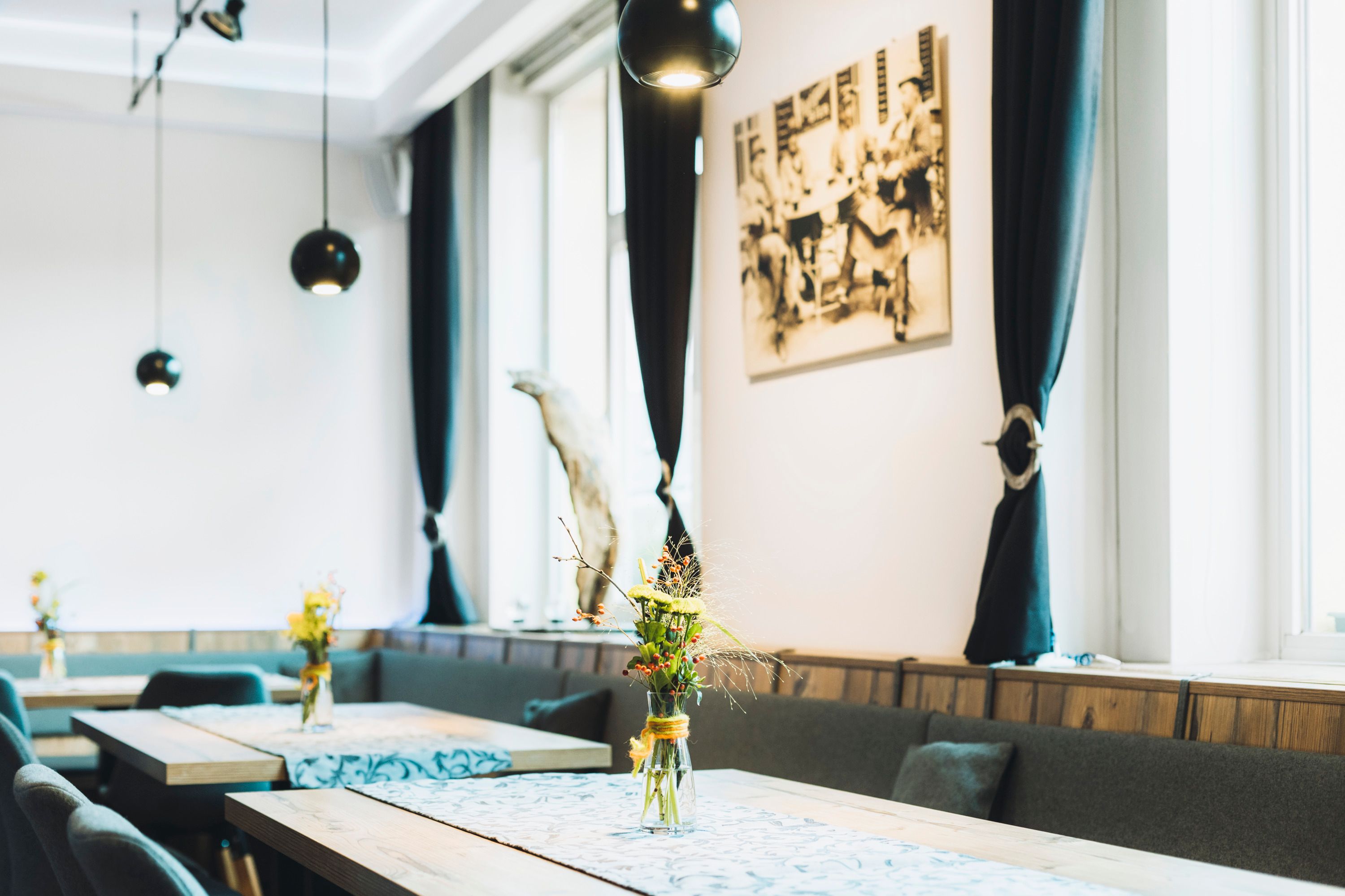 Egy elegáns kávézó belső nézete, fa asztalokkal, virágvázákkal és fekete függönyökkel.