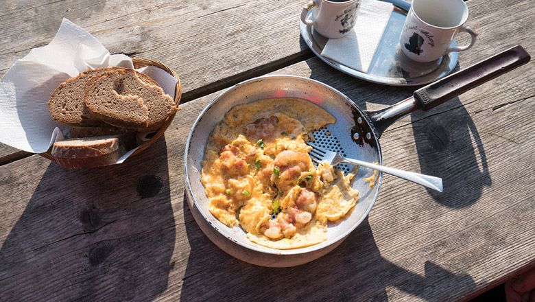 Rántottát és szalonnát tartalmazó serpenyő a faasztalon, mellette kenyérkosár és két csésze.