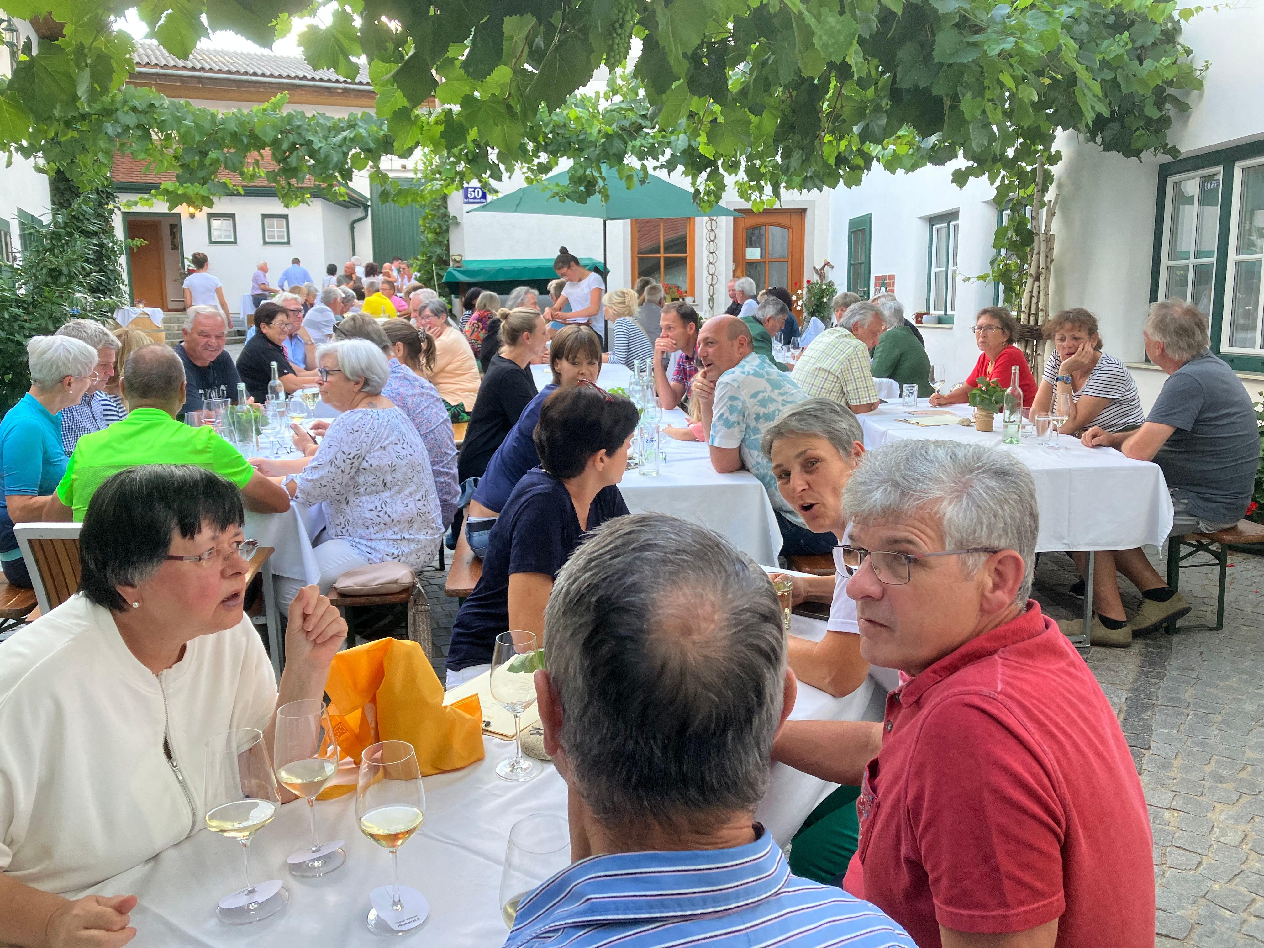 Az emberek kint ülnek az asztaloknál a szőlőtőkék alatt és beszélgetnek.