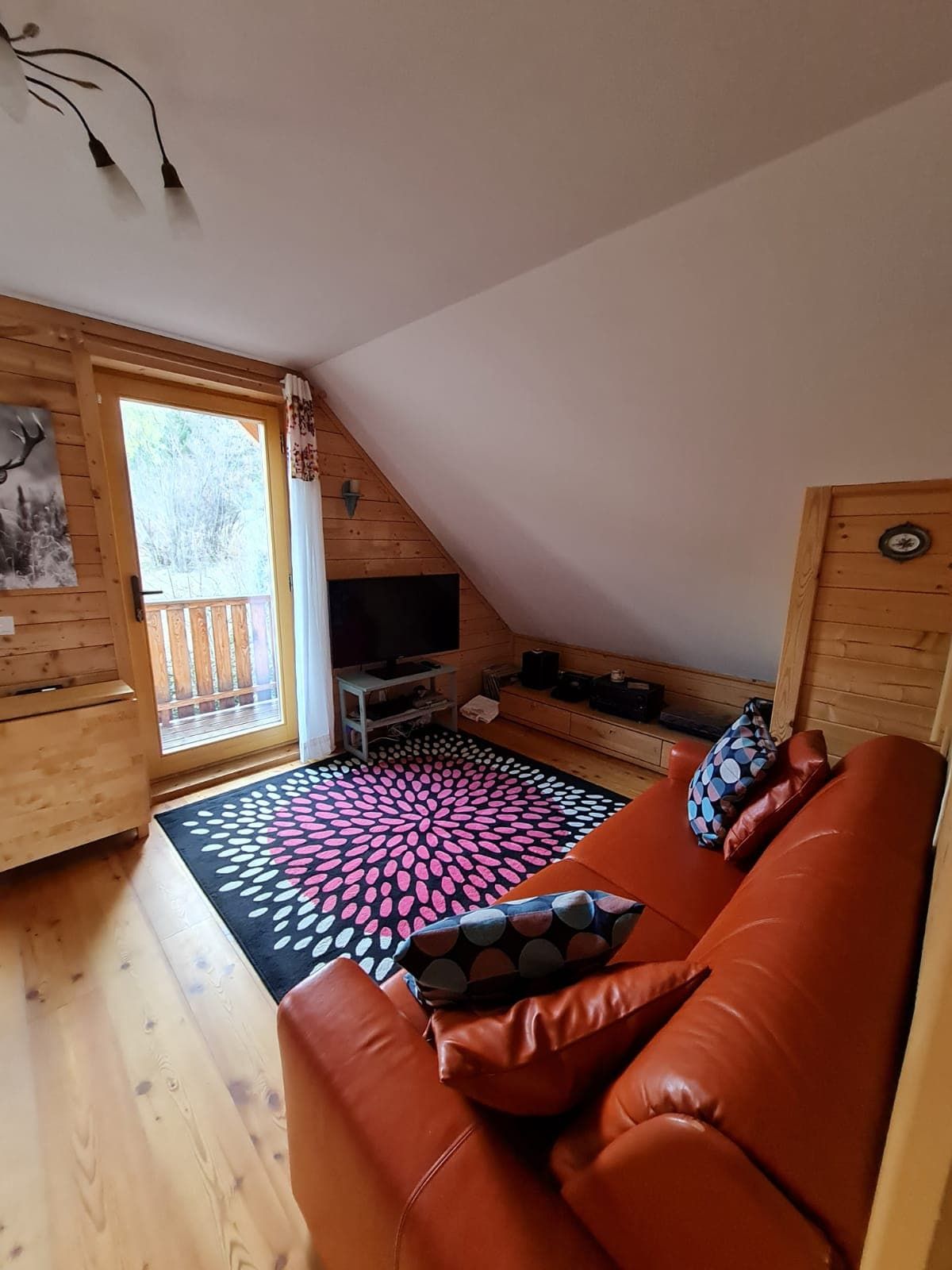 Hangulatos nappali piros kanapéval, fából készült falakkal és színes szőnyeggel az erkélyablak előtt.