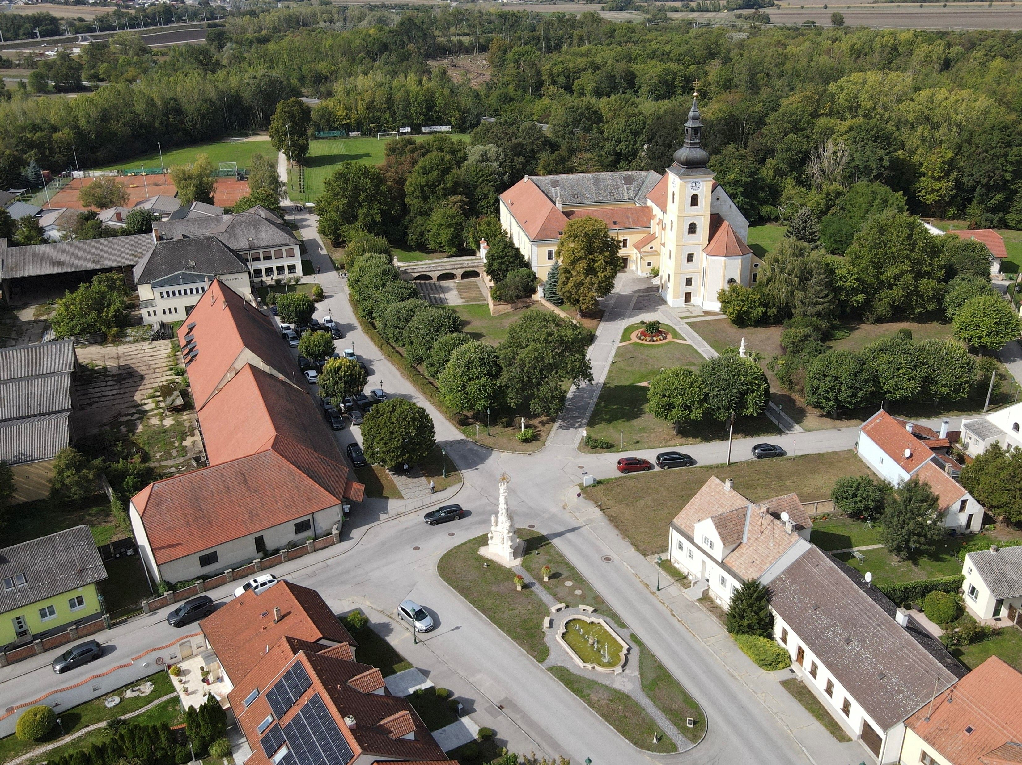Weikendorf légi felvétele a templommal, a falu térrel, a szökőkúttal és a környező házakkal egy erdőszélén.