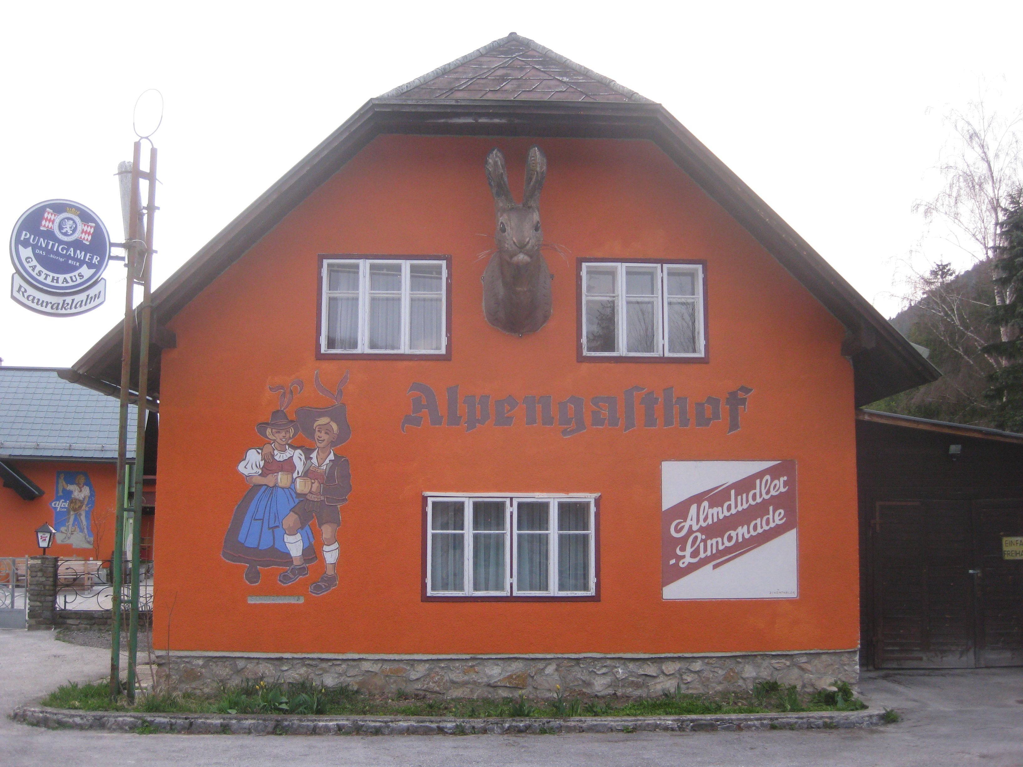 Narancsszínűre festett épület "Alpengasthof" felirattal, táncoló párt ábrázoló falfestmény és Almdudler-reklám.