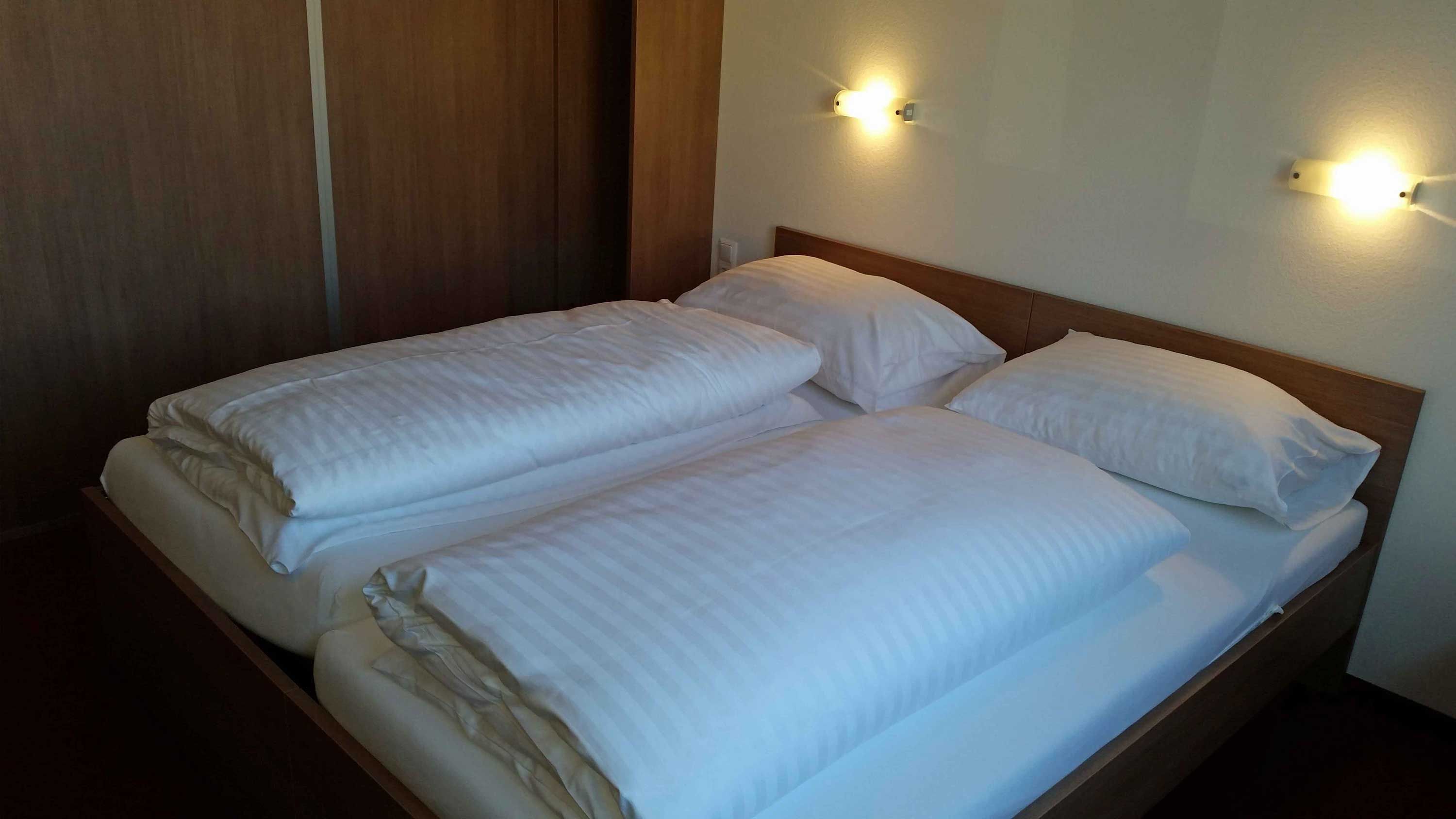 Kétágyas ágy egy motelszobában, fehér paplanokkal és párnákkal, két fali lámpa megvilágításában.