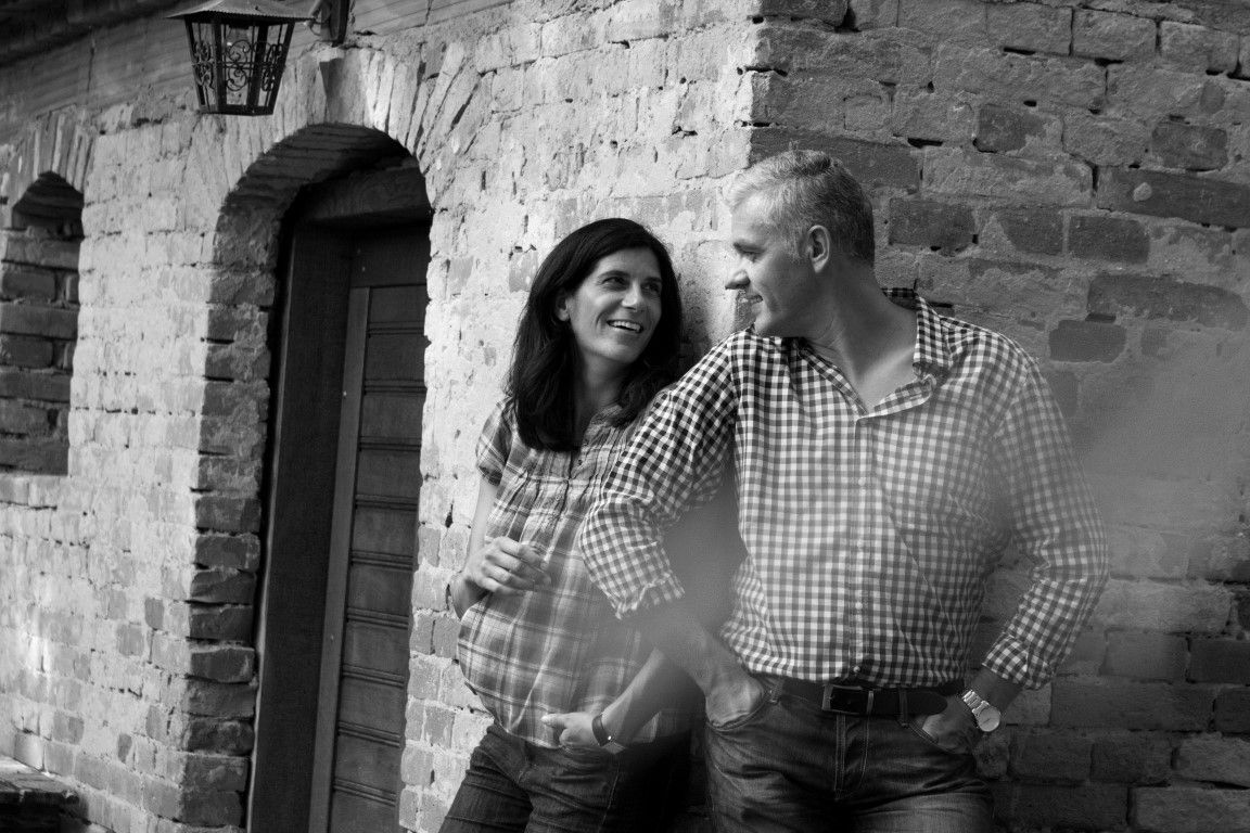 Egy férfi és egy nő mosolyogva támaszkodik egy téglafalnak.