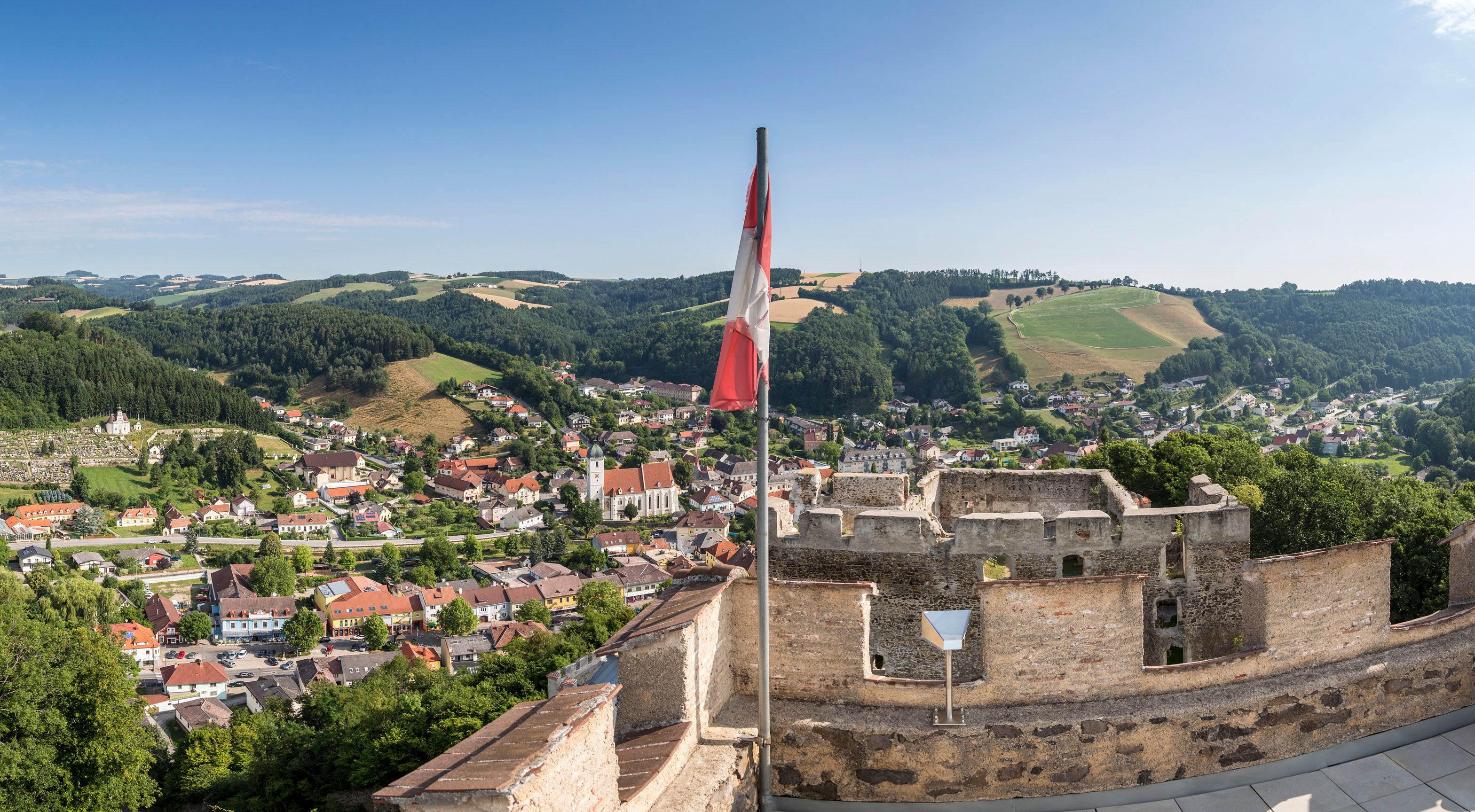 Panorámakép egy romos várból egy városra, a háttérben dombokkal, az előtérben osztrák zászlóval.