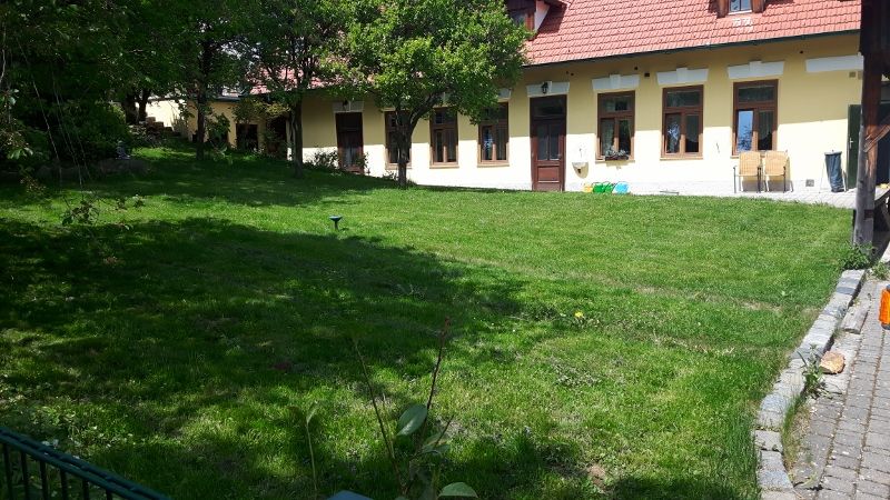 Zöld kert egy sárga épület előtt, piros tetőcseréppel és több ablakkal.