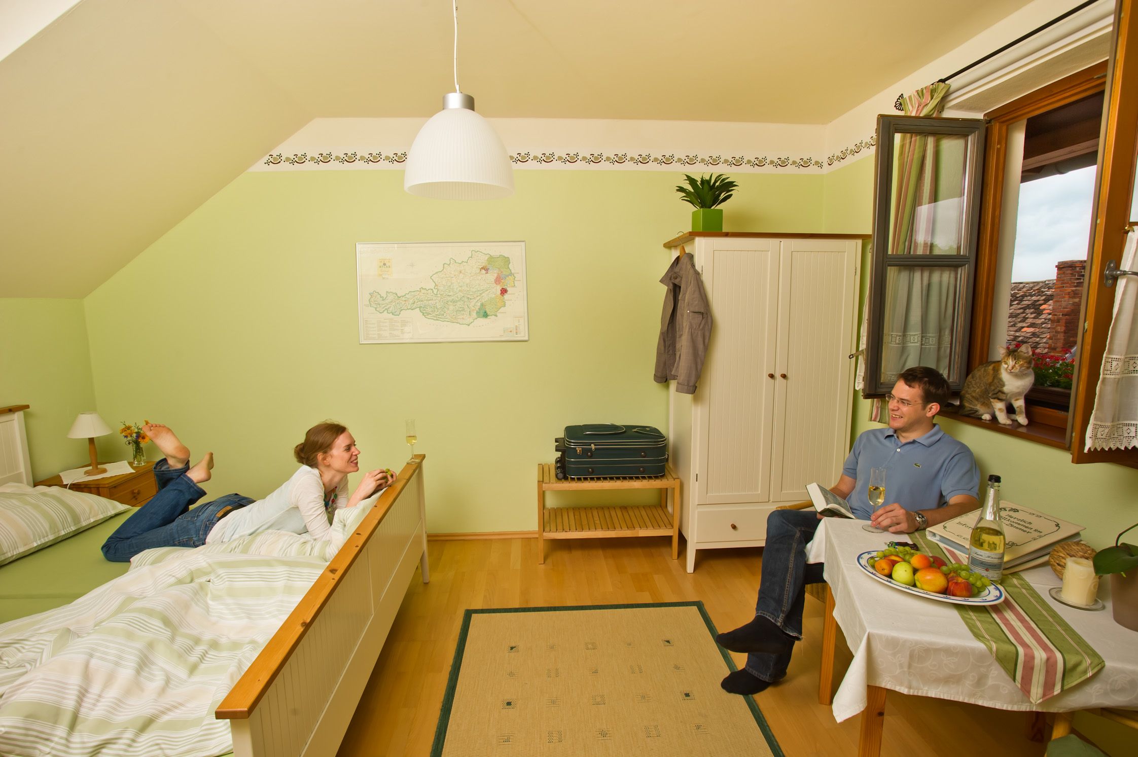 Hangulatos vendégszoba, ahol egy pár pihen. Egy nő fekszik az ágyon, egy férfi ül az asztalnál. Az ablaknál egy macska ül.