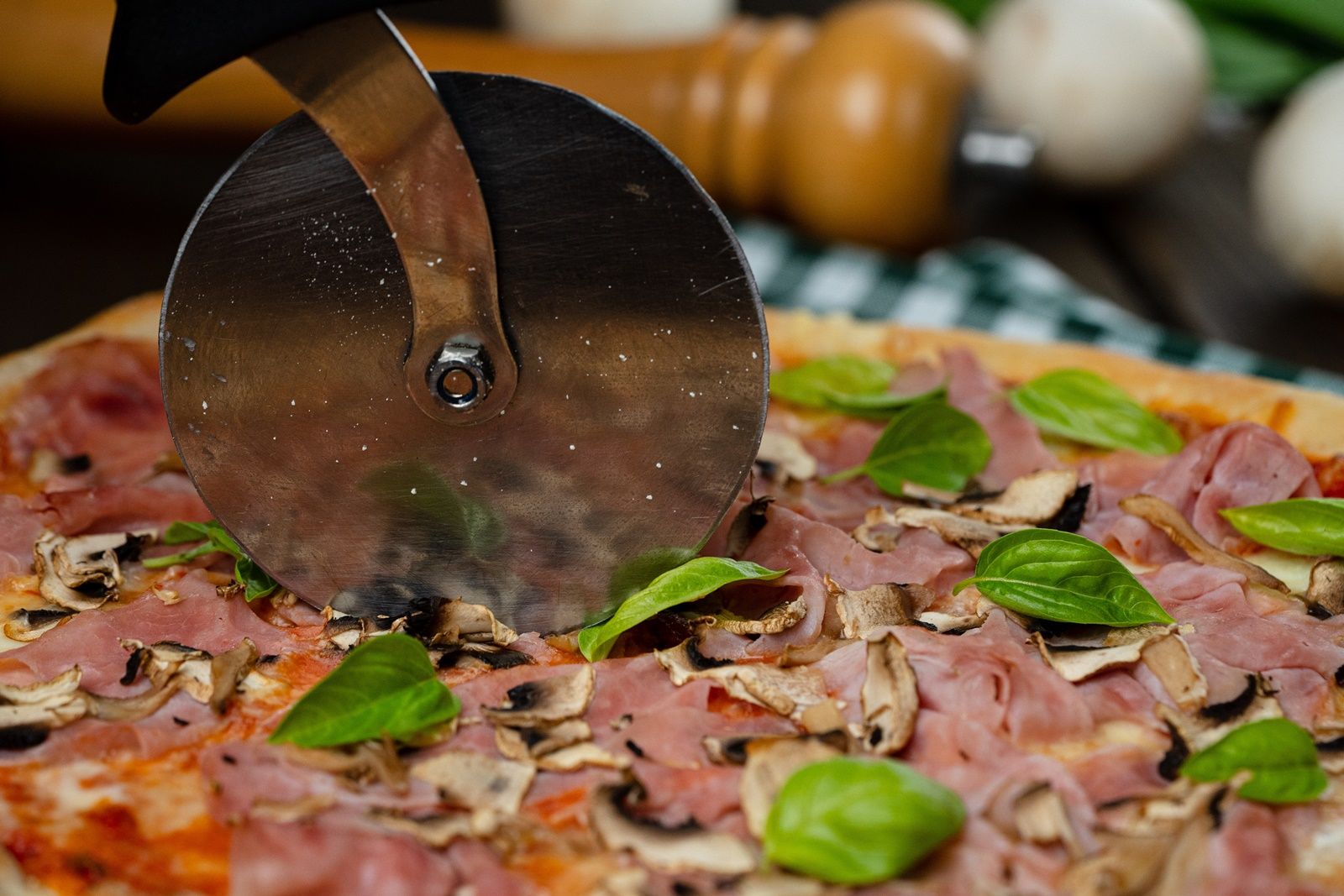 Közelkép egy sonkás, gombás és bazsalikomos pizzáról, amelyet pizzavágóval vágnak.
