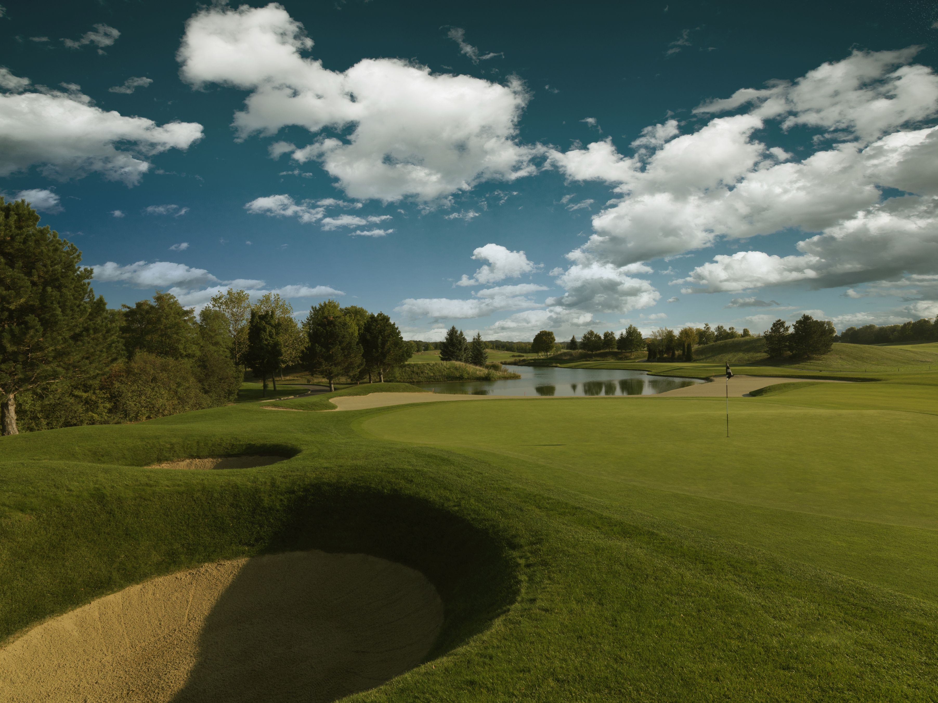 Golfpálya homokbunkerrel, zölddel és tóval, kék felhős égbolt alatt.