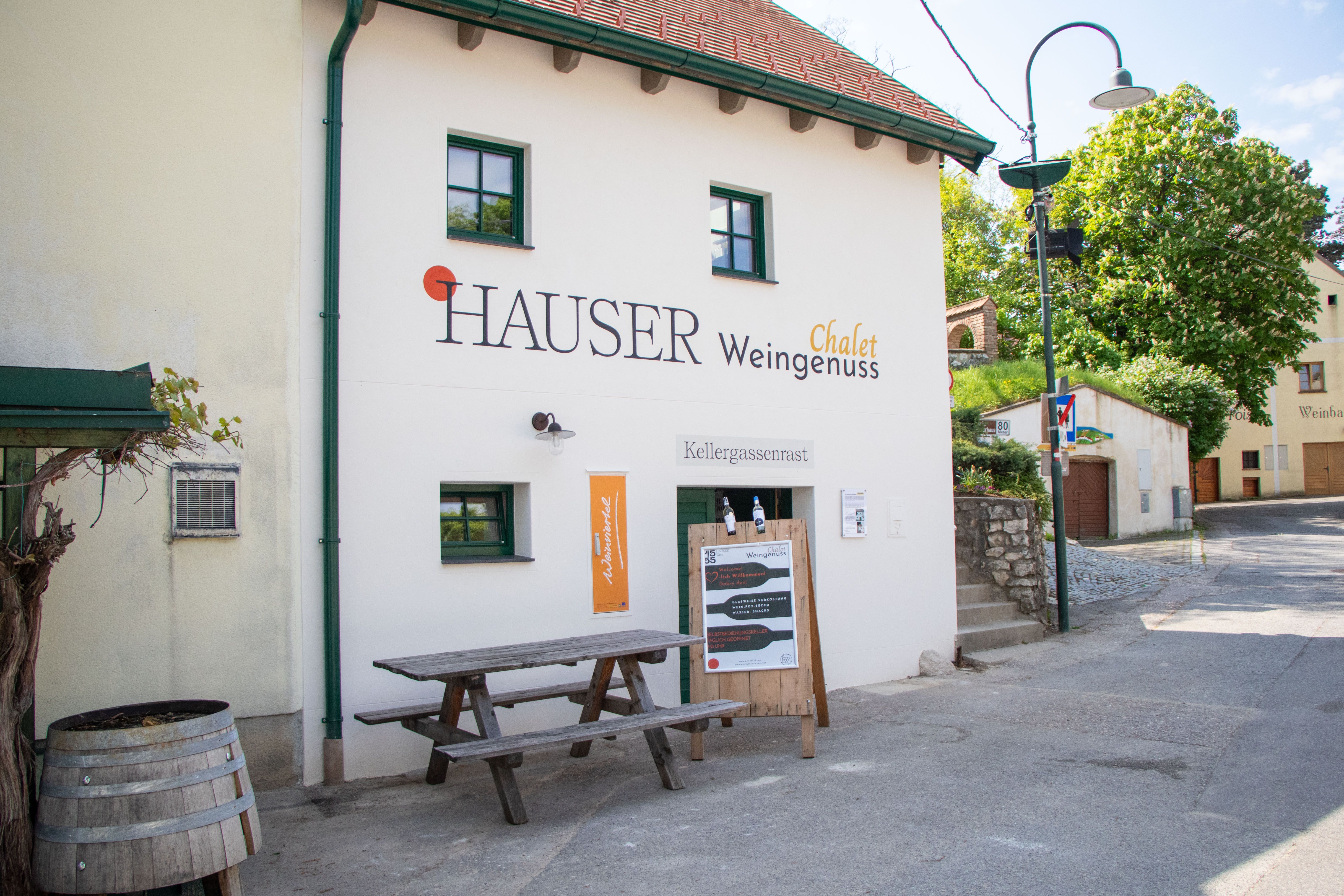 A "Hauser Chalet Weingenuss" feliratú épület külső nézete. Az épület előtt egy faasztal és egy boroshordó áll.