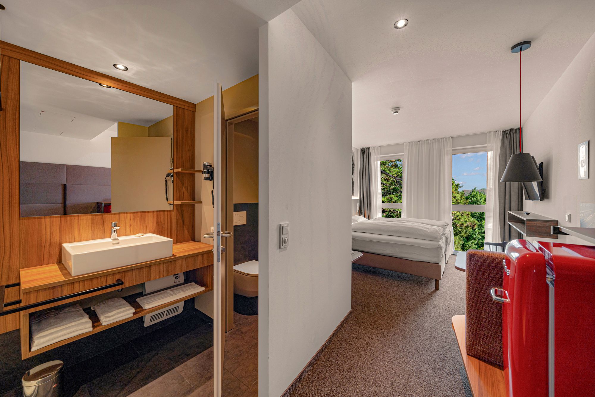 Egy modern szállodai szoba belső nézete fürdőszobával, ággyal és piros hűtőszekrénnyel.