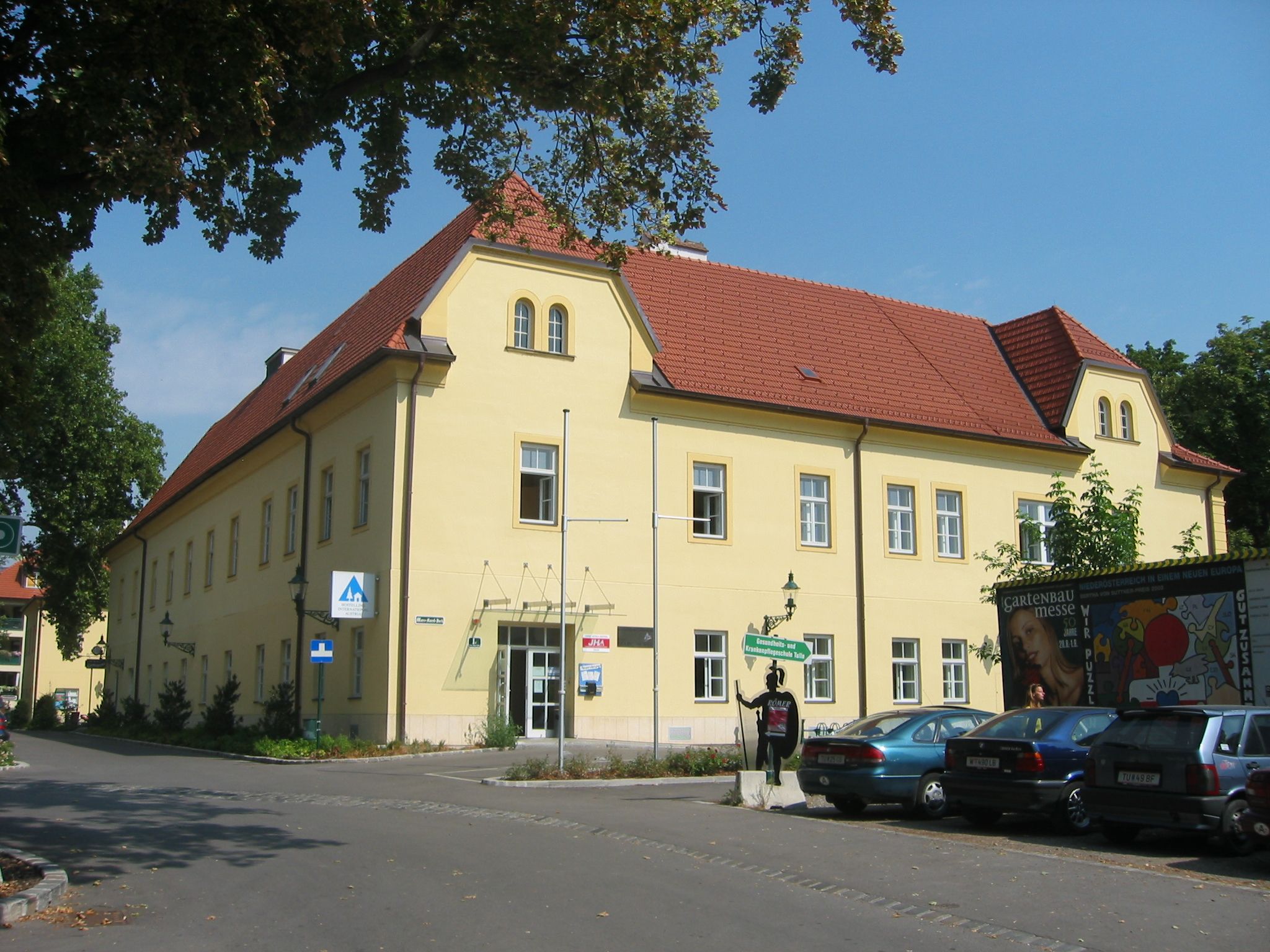 Külső nézet egy sárga, piros tetejű épületről, a Junges Hotel Tullnról, amelyet fák és parkoló autók vesznek körül.