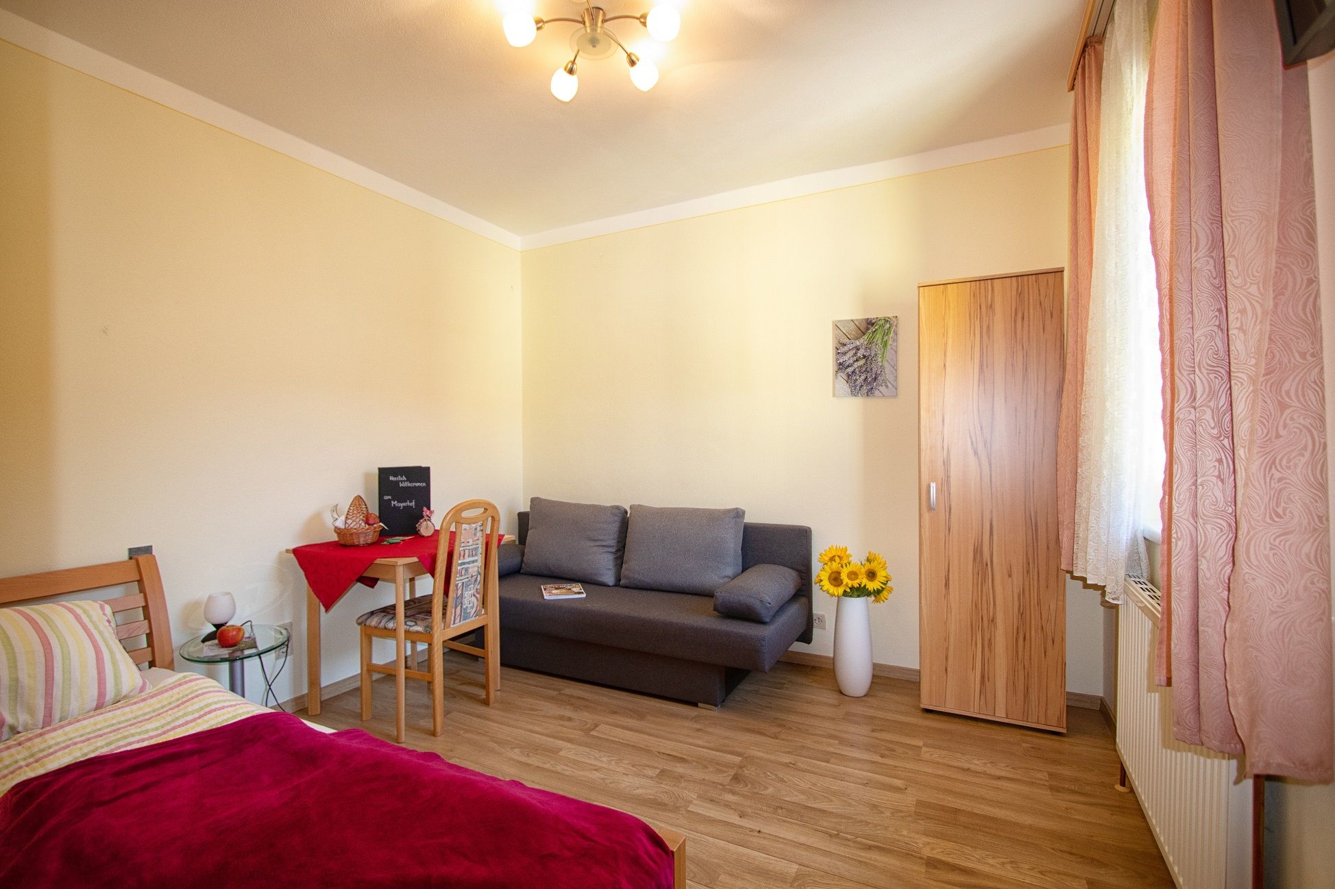 Hangulatos szoba ággyal, kanapéval, asztallal és szekrénnyel, napraforgókkal és egy képpel a falon.