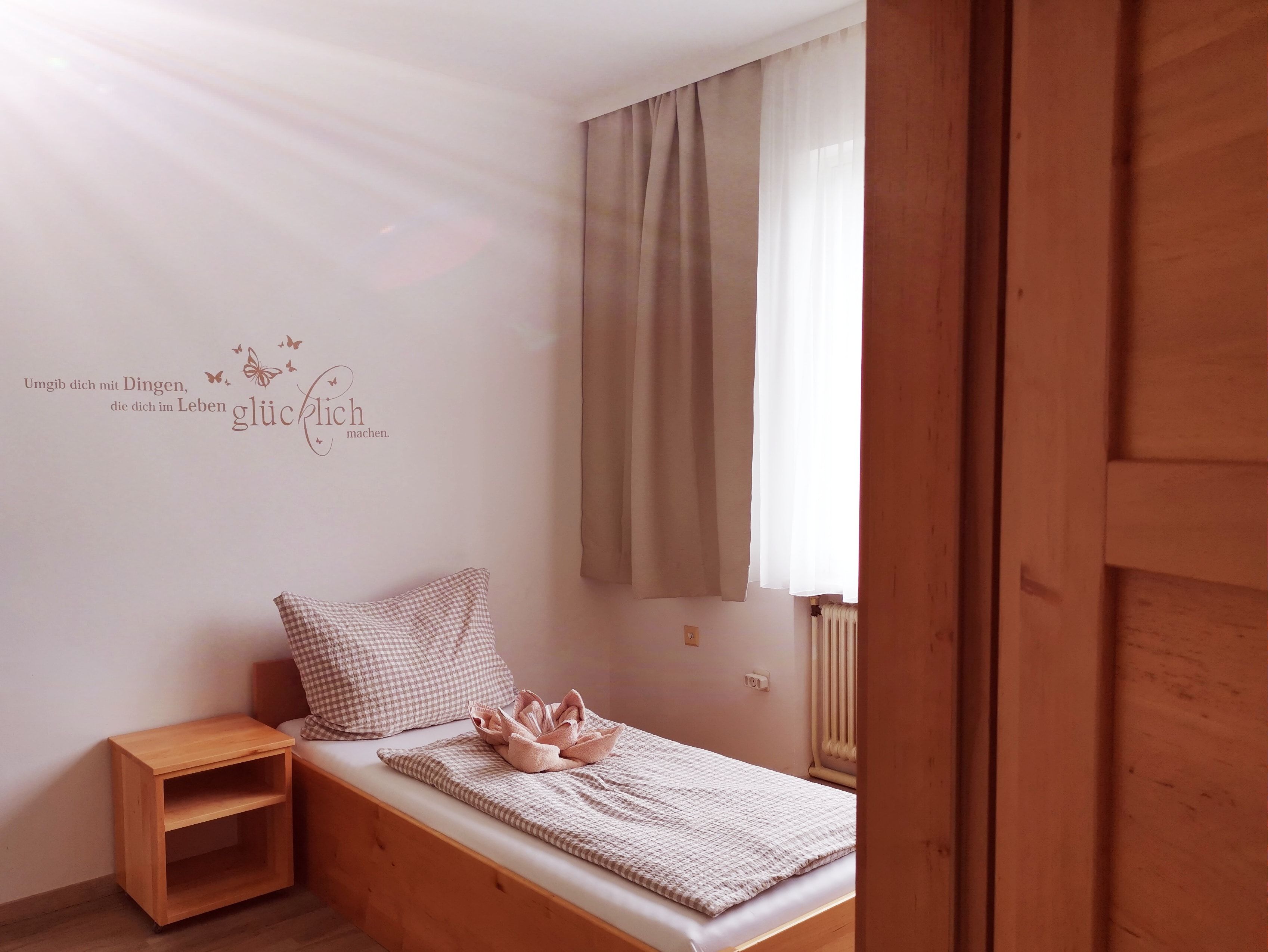 Egyszerű vendégszoba egy egyszemélyes ággyal, fa polcokkal és egy idézettel a falon.