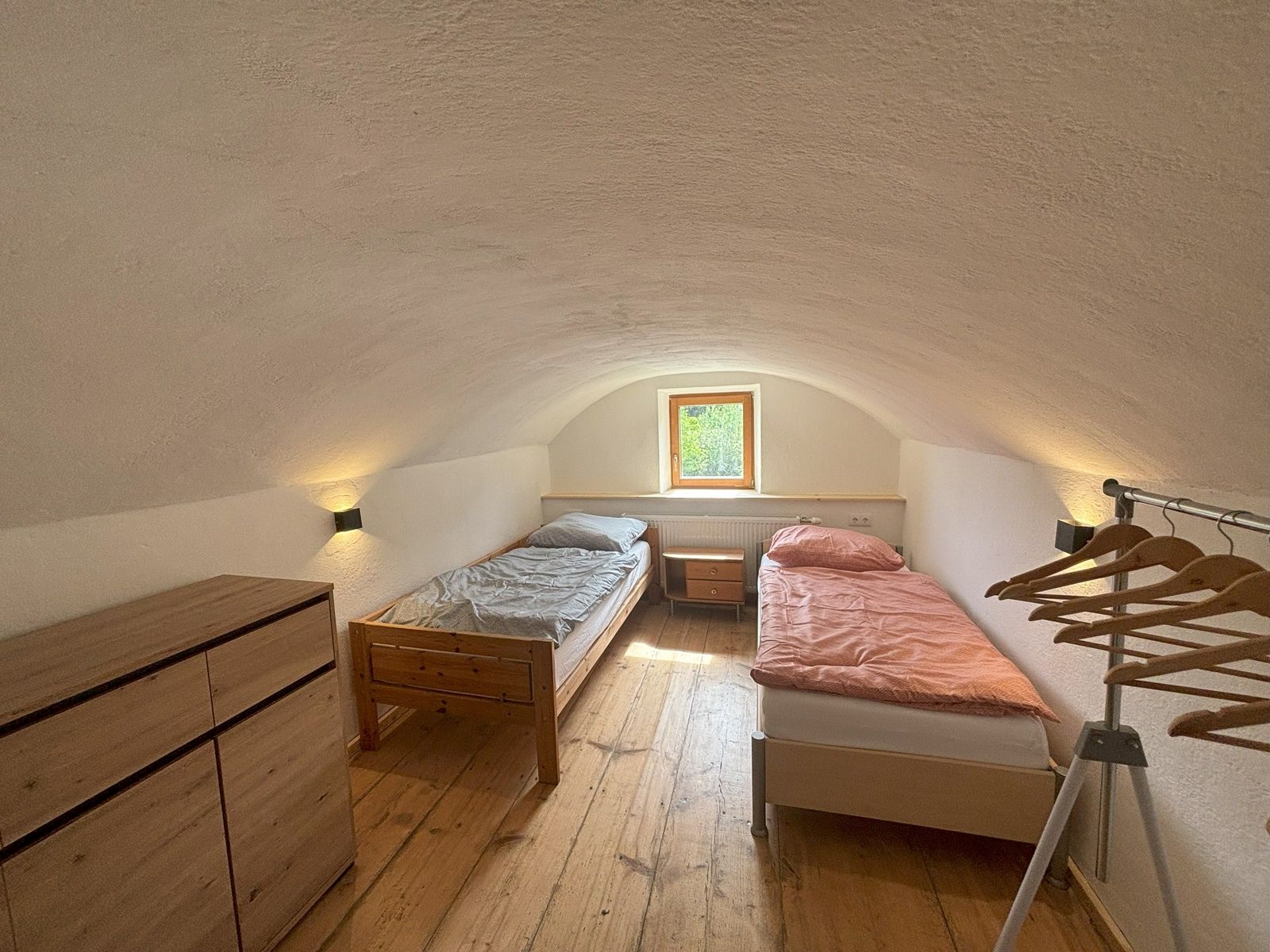 Egyszerű kétágyas szoba két egyszemélyes ággyal, komóddal és ruhatartóval. Egy kis ablakon keresztül természetes fény szűrődik be.