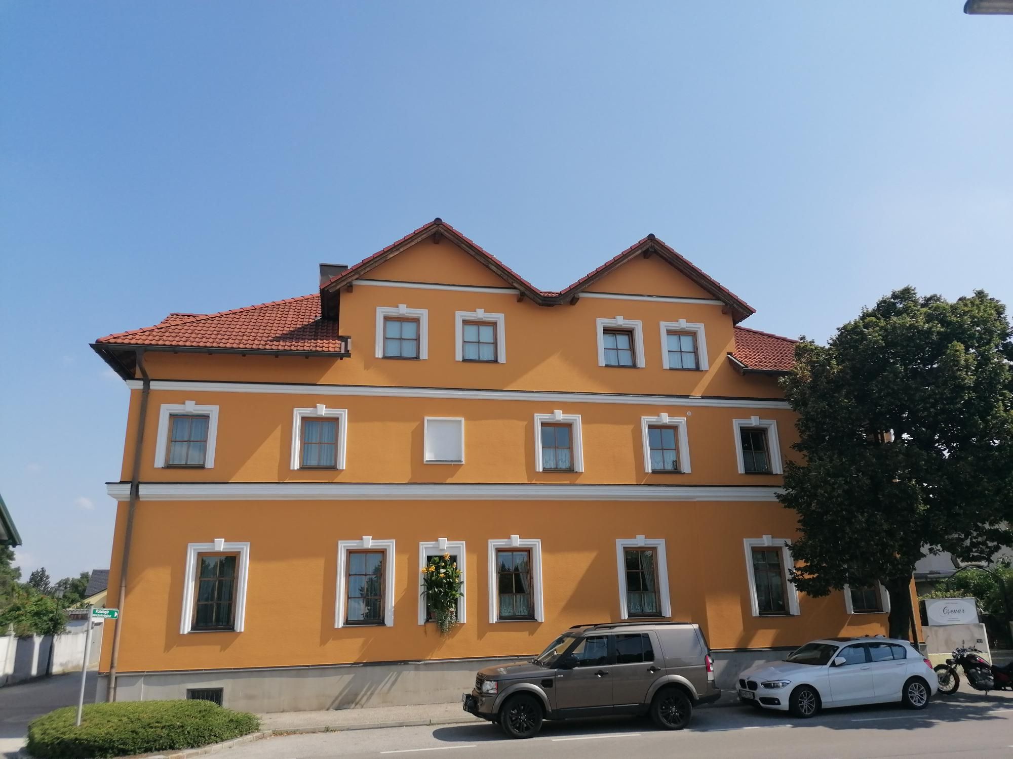 Sárga épület piros tetőcseréppel és több ablakkal, előtte parkoló autók és egy fa.