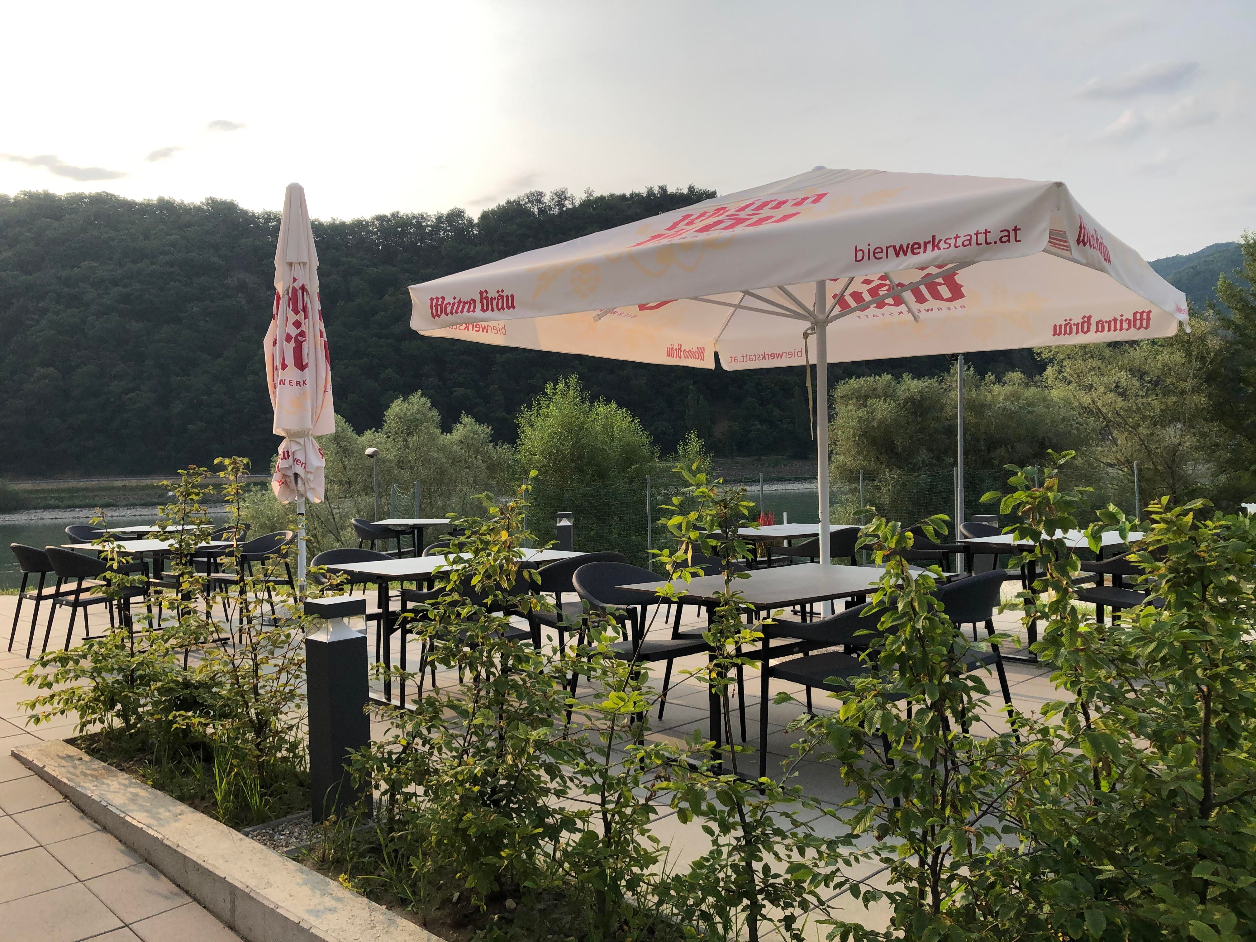 Terasz asztalokkal, székekkel és napernyőkkel, a természet által körülvéve.