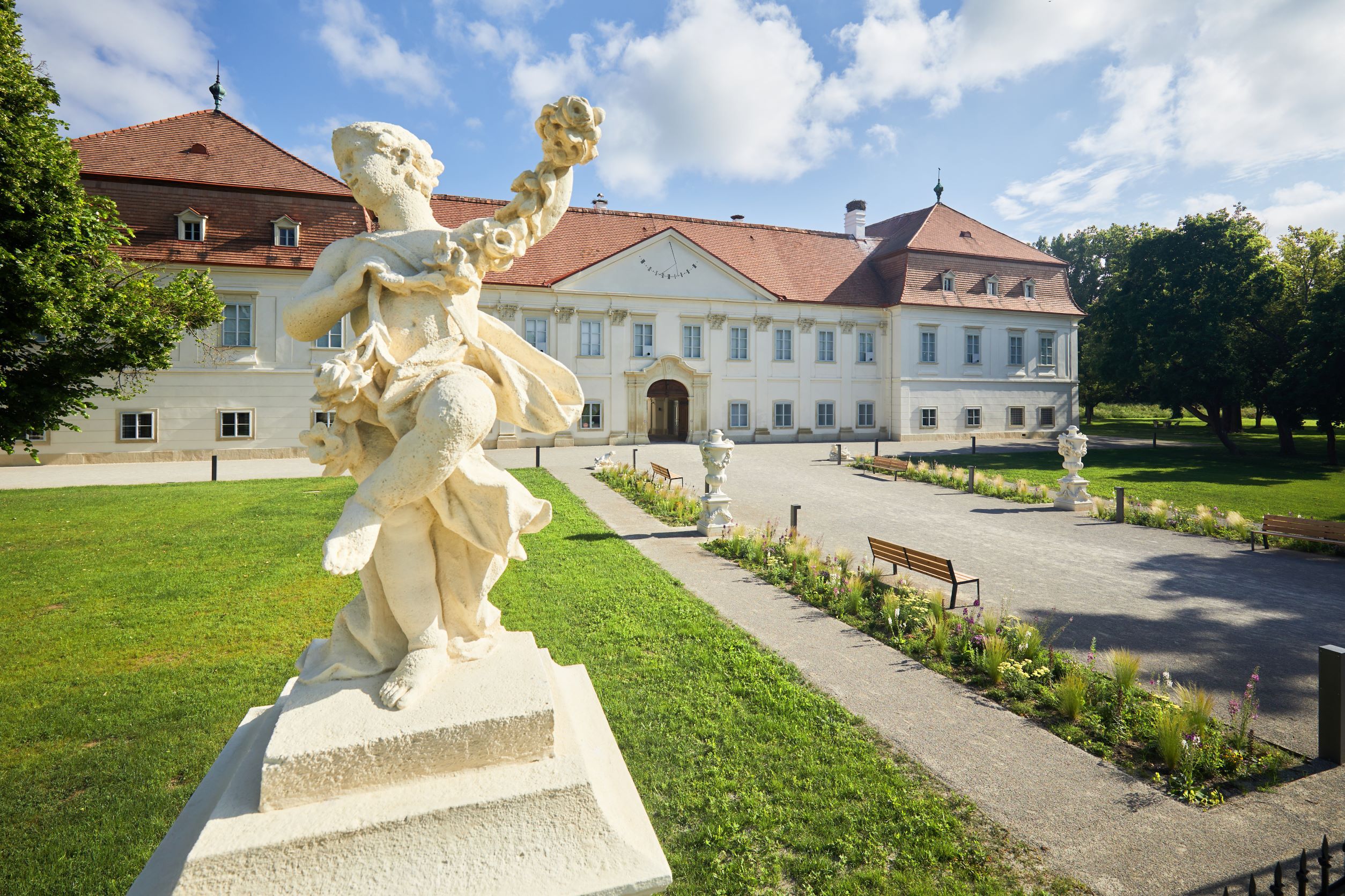 Barokk szobor egy vörös tetővel és gondozott kerttel rendelkező történelmi épület előtt.