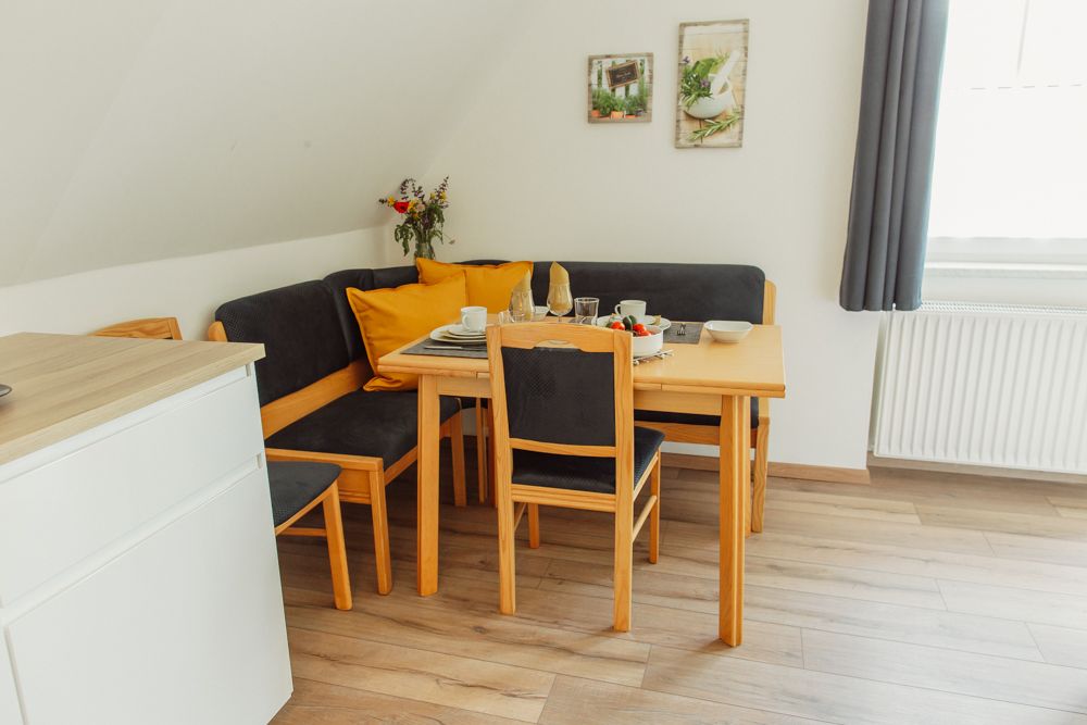 Étkezőhelyiség fa asztallal, székekkel és sarokpaddal, étkészlettel, virágcsokorral és dekorációval a falon.