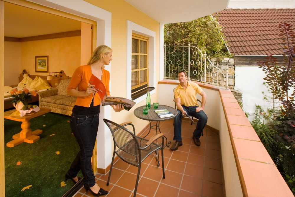 Egy férfi ül az erkélyen, egy nő jön ki a szobából egy magazinnal. Asztal italokkal, hangulatos légkör.