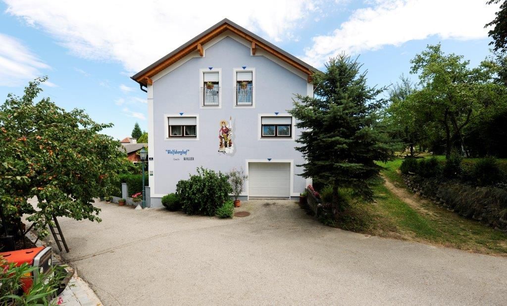 Kétszintes ház, kék homlokzattal és fatetővel, fákkal és kövezett úttal körülvéve.