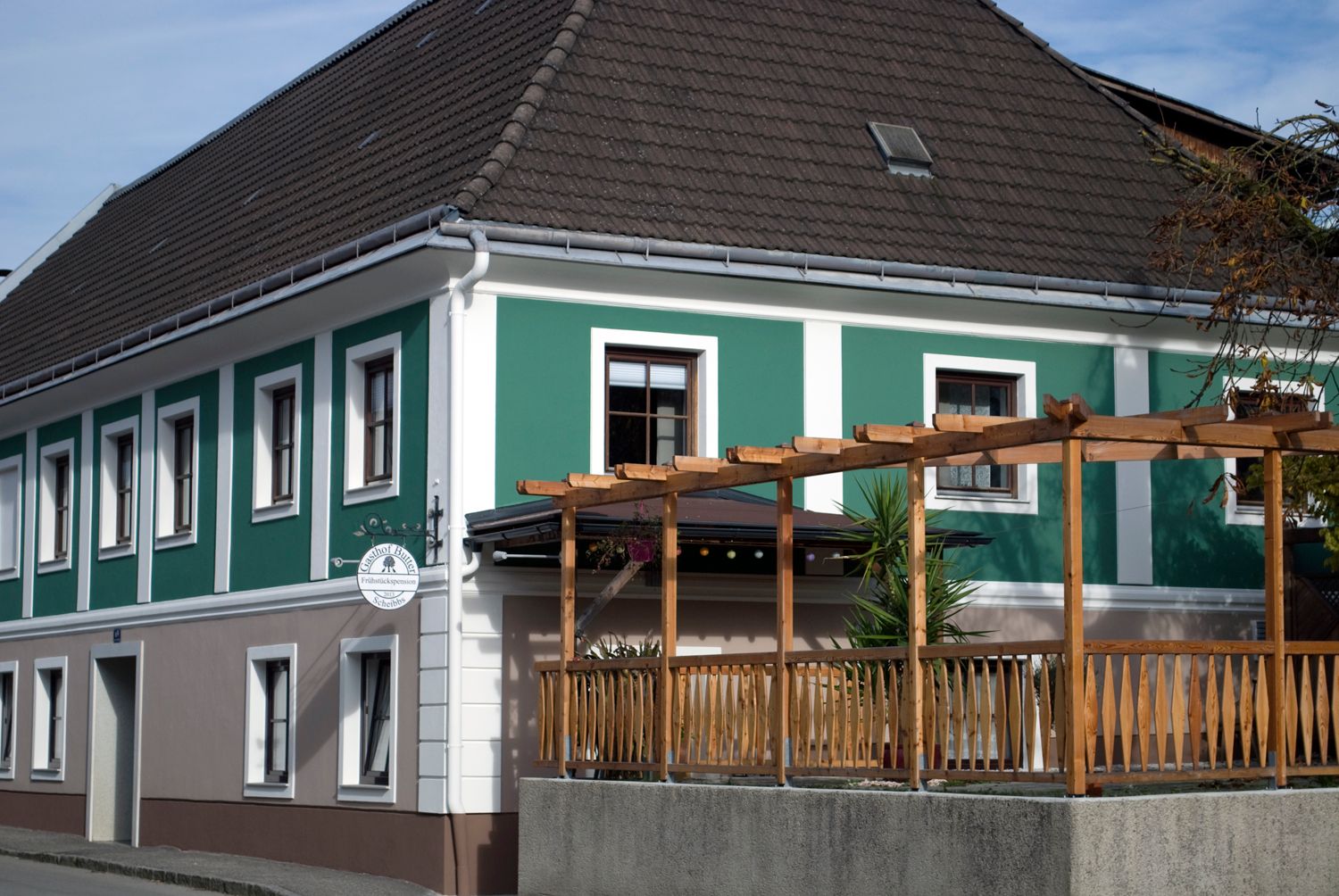 Egy panzió külső nézete zöld-fehér homlokzattal és fa pergolával.