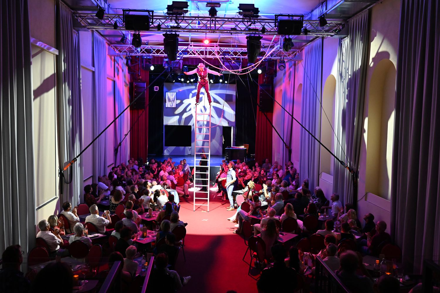 Egy művész egy létrán egyensúlyoz egy zsúfolt színházteremben, színes világítással.