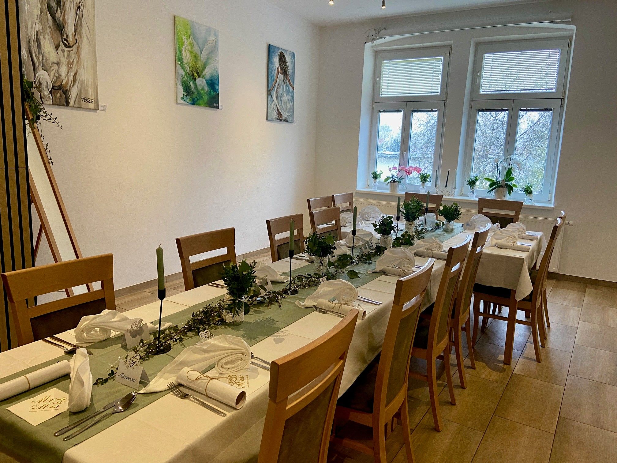 Ünnepélyesen megterített asztal egy szobában, ahol a falon műalkotások és nagy ablakok vannak.