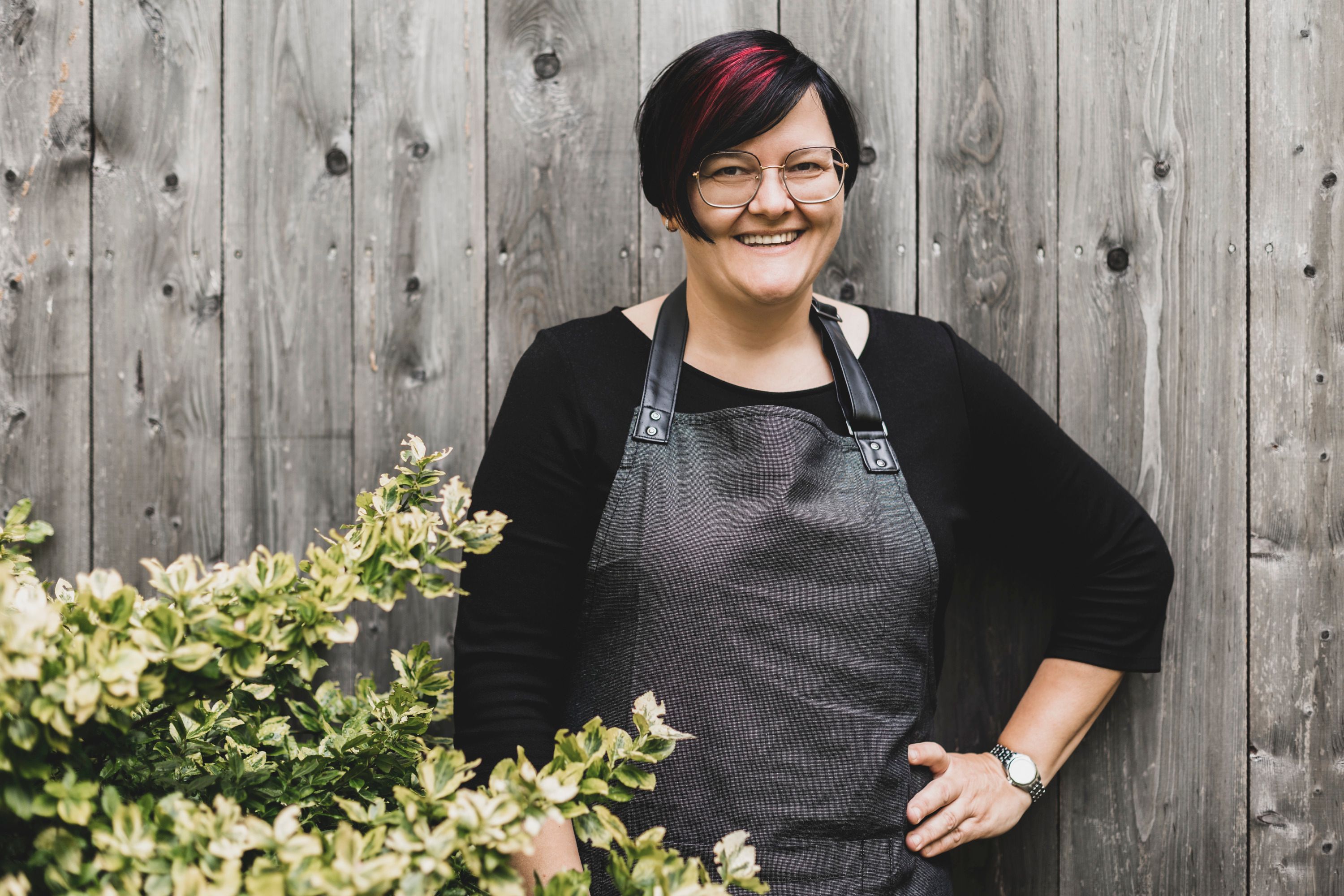 Egy szemüveges, kötényes nő áll mosolyogva egy bokor melletti fal előtt.