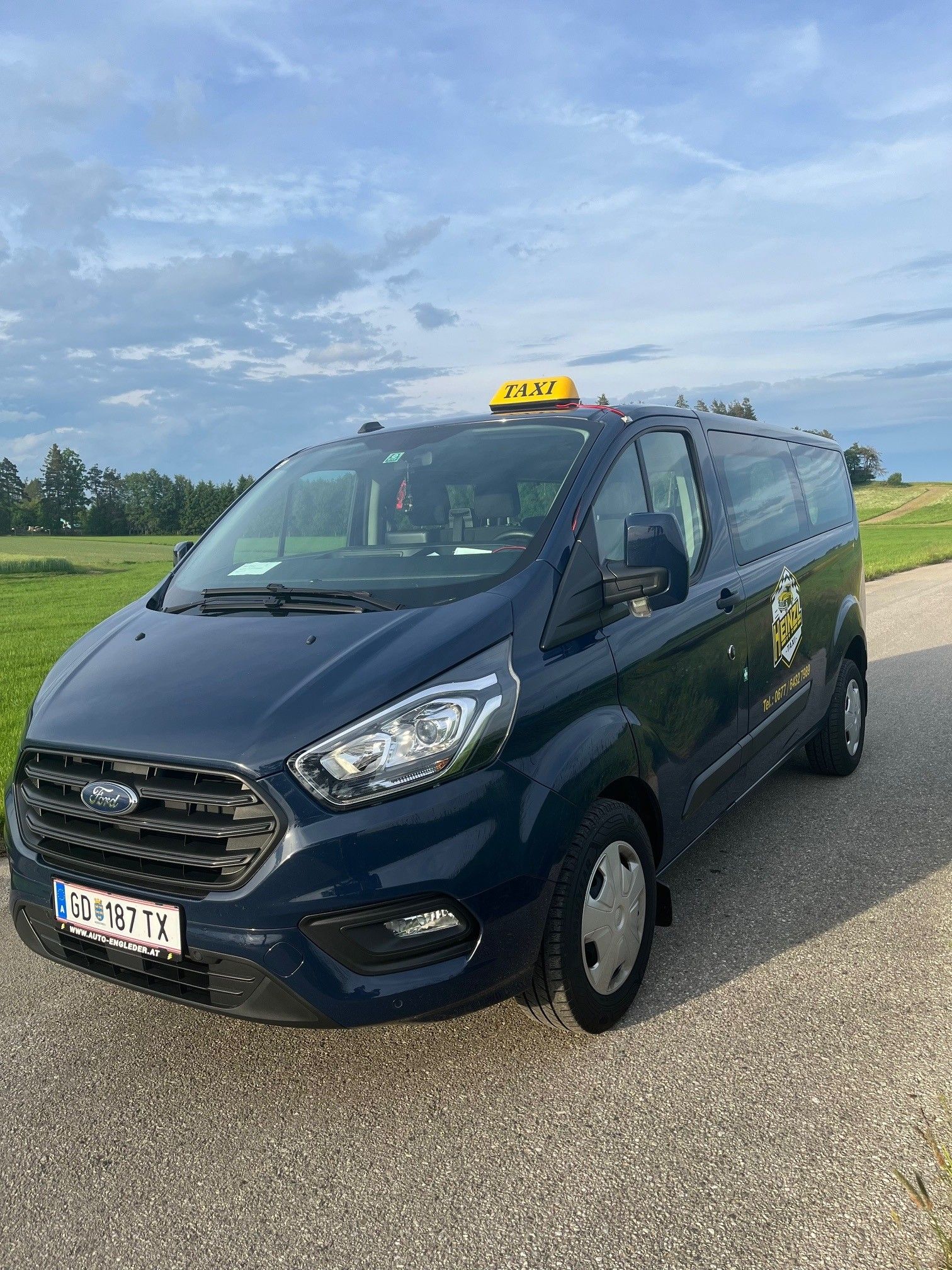 Egy kék Ford taxi parkol egy országúton, zöld rét és felhős égbolt előtt.