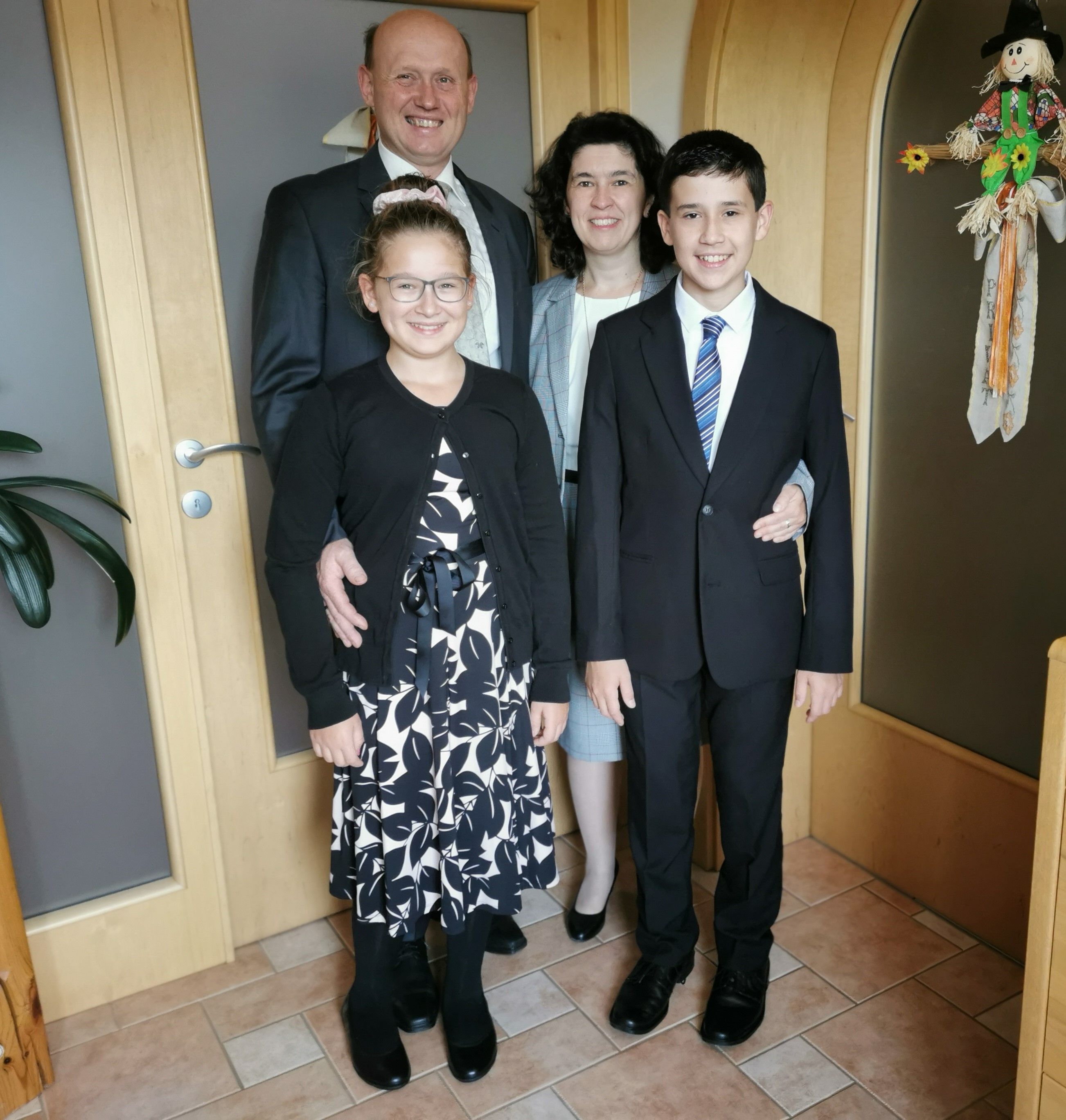 Egy hivatalos ruhába öltözött család áll mosolyogva a folyosón.