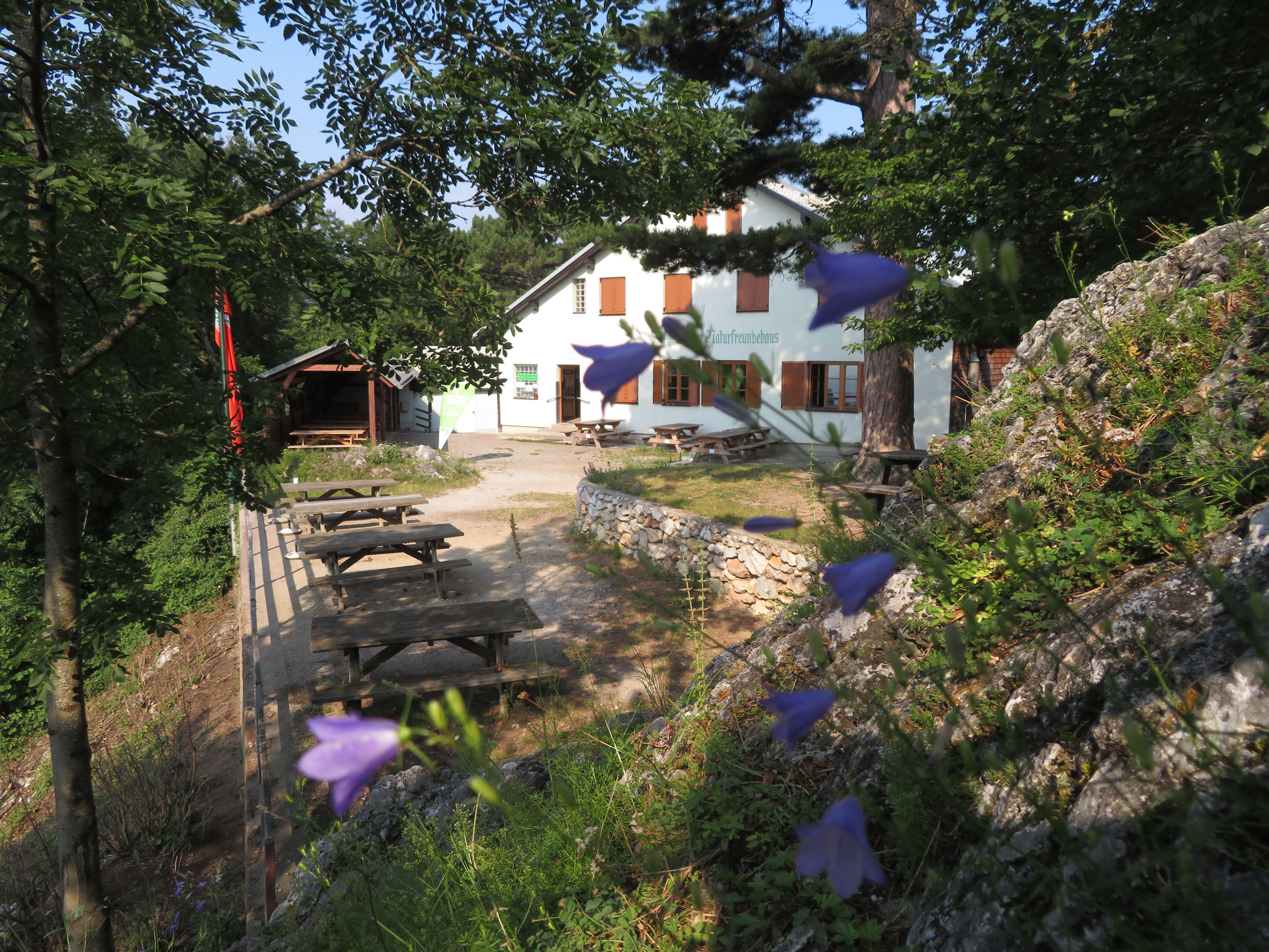 Egy fehér, "Naturfreundehaus" feliratú épület, amelyet fák és virágok vesznek körül. Az előtérben fapadok és asztalok láthatók.