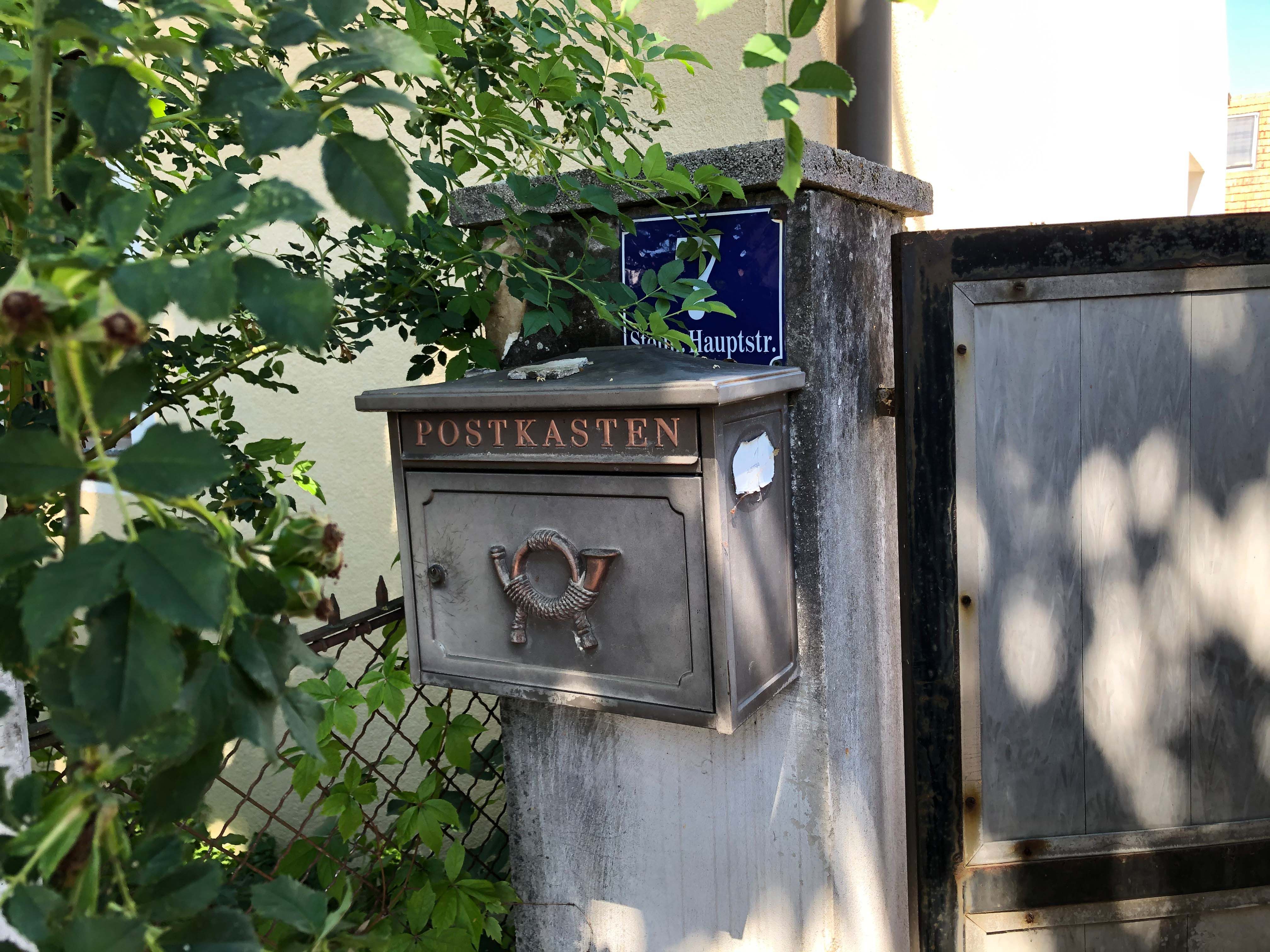 Egy régi postaláda "Postbox" felirattal egy kapu melletti falon, növényekkel körülvéve.