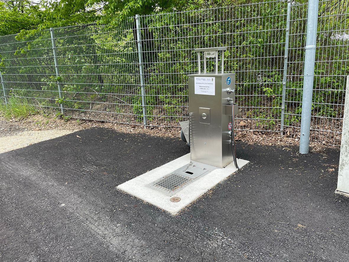 Fém vízellátó állomás egy kerítés mellett az aszfalton.