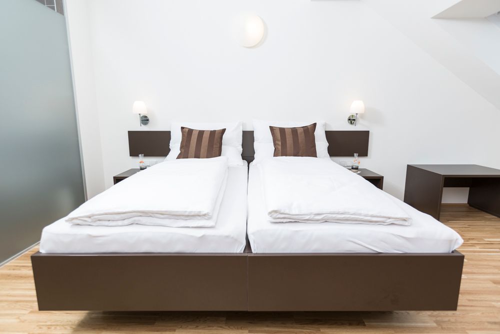 Modern vendégszoba két egyszemélyes ággyal, fehér paplanokkal és barna párnákkal, fapadlóval és éjjeliszekrényekkel, lámpákkal.