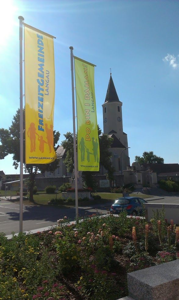 Két színes zászló "Freizeitgemeinde Langau" és "Do bin i daham" felirattal egy templom előtt.