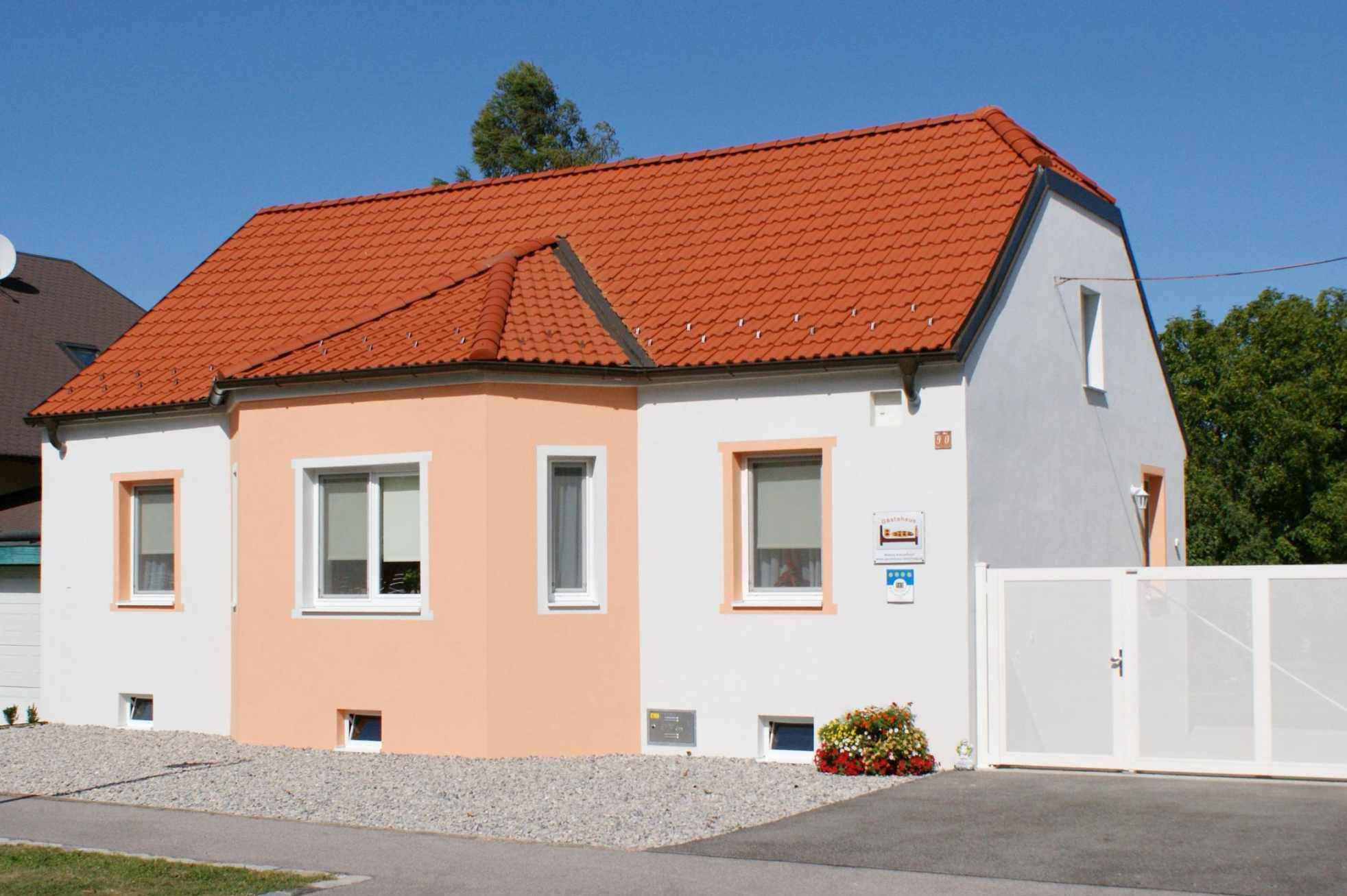 Egy kis, egyemeletes ház, narancssárga színű tetővel és fehér homlokzattal, kék ég alatt.