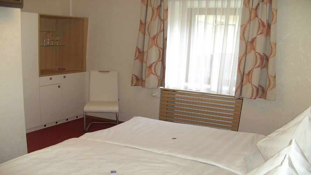 Egy szállodai szoba egy ággyal, egy székkel, egy szekrénnyel és egy függönyös ablakkal.