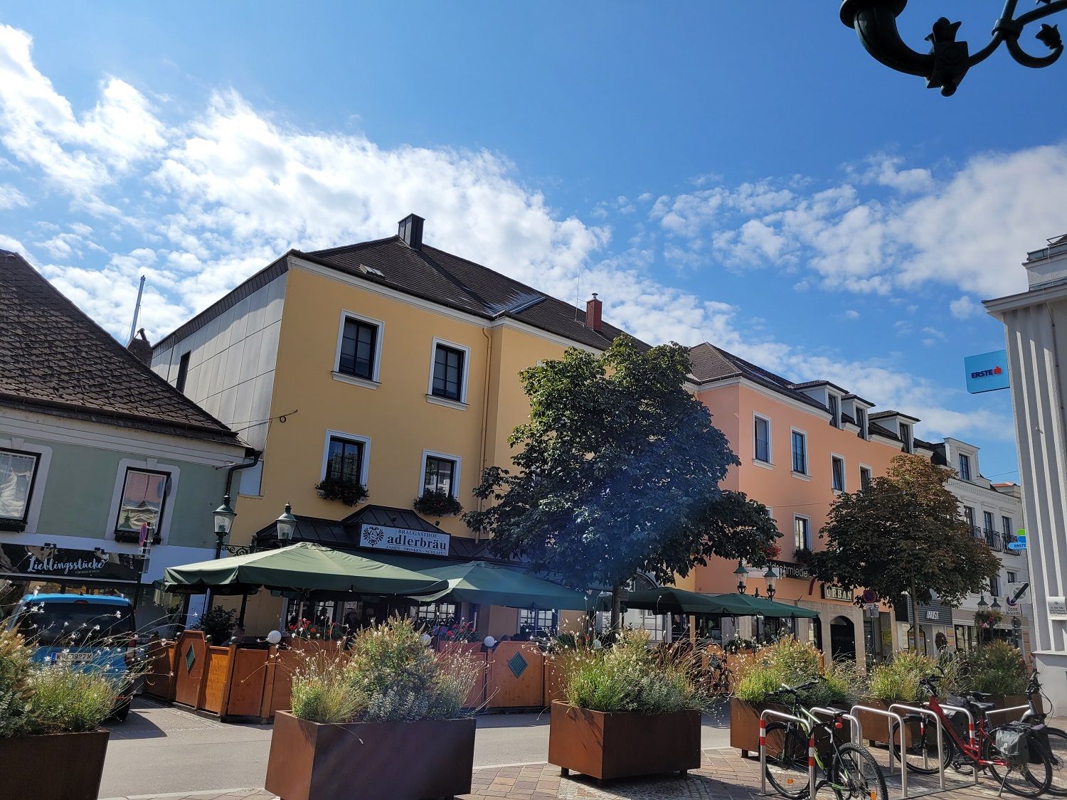Utcai jelenet sárga épülettel, kávézó napernyőkkel és kerékpárokkal az előtérben.