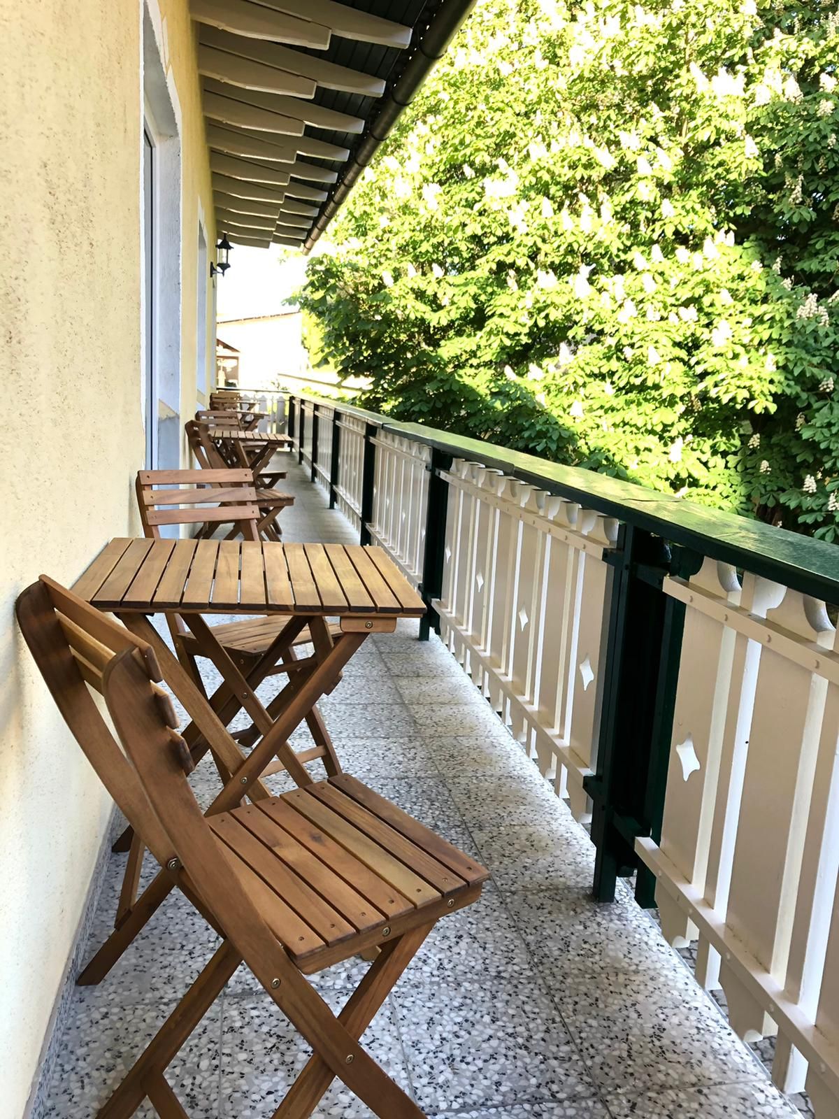 Fából készült erkély asztallal és székekkel, mellette zöld fák.