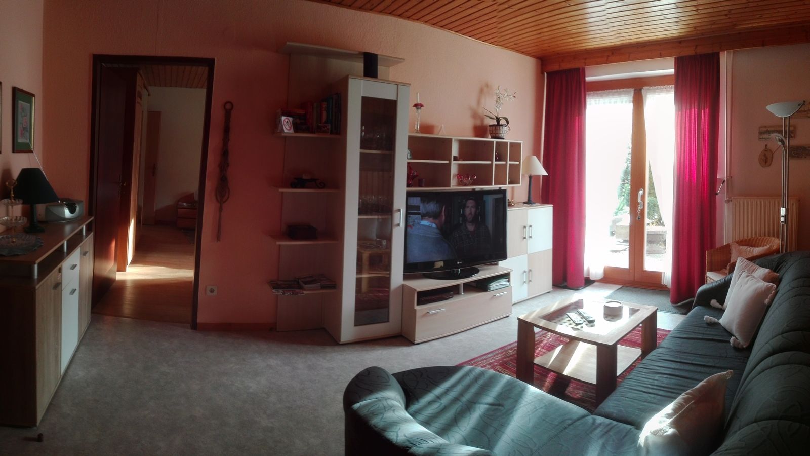 Hangulatos nappali kanapéval, TV-vel, polccal és üvegajtóval, piros függönyökkel.