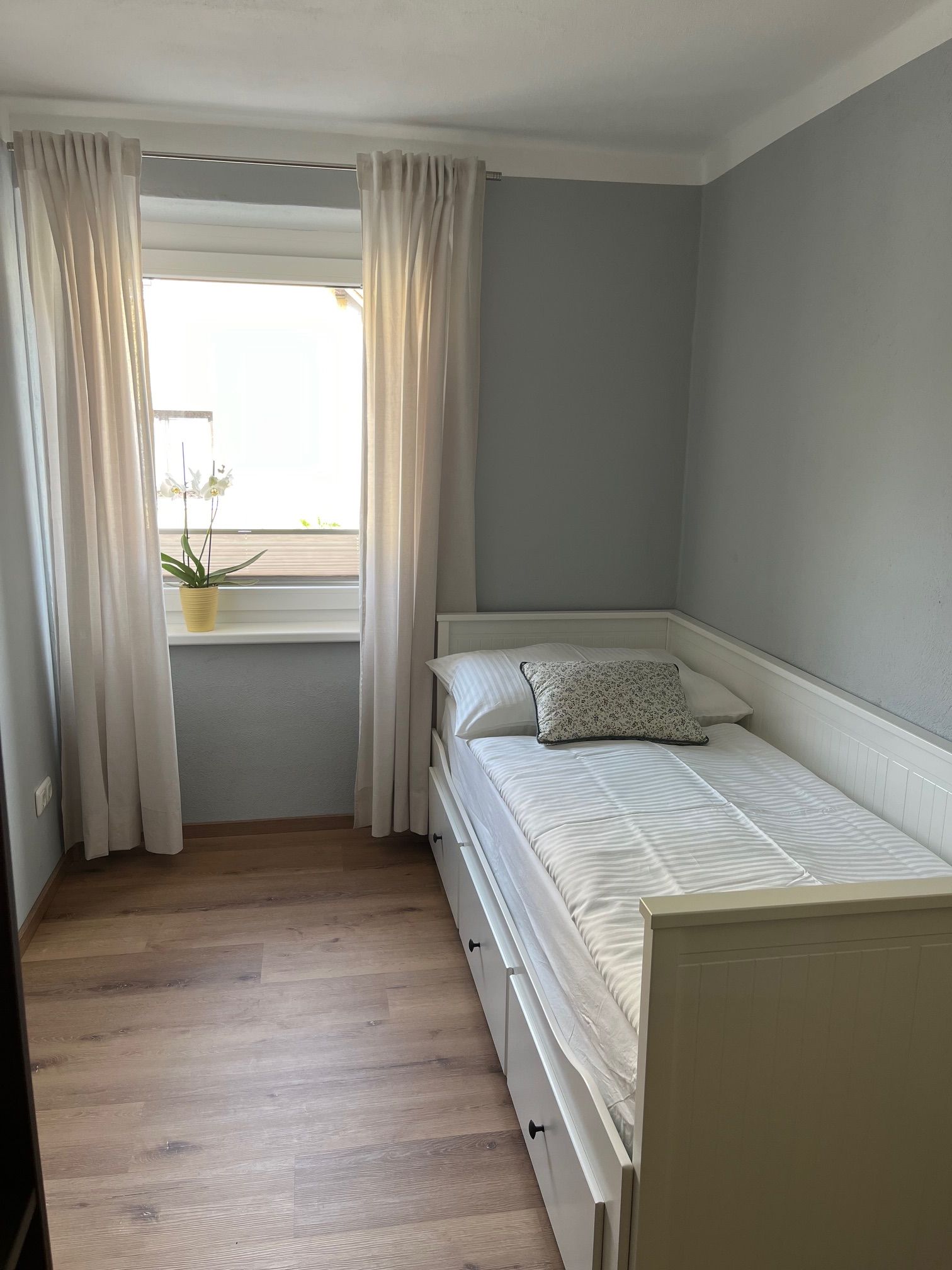 Kis hálószoba egyszemélyes ággyal, fehér függönyökkel és egy növénnyel az ablakpárkányon.