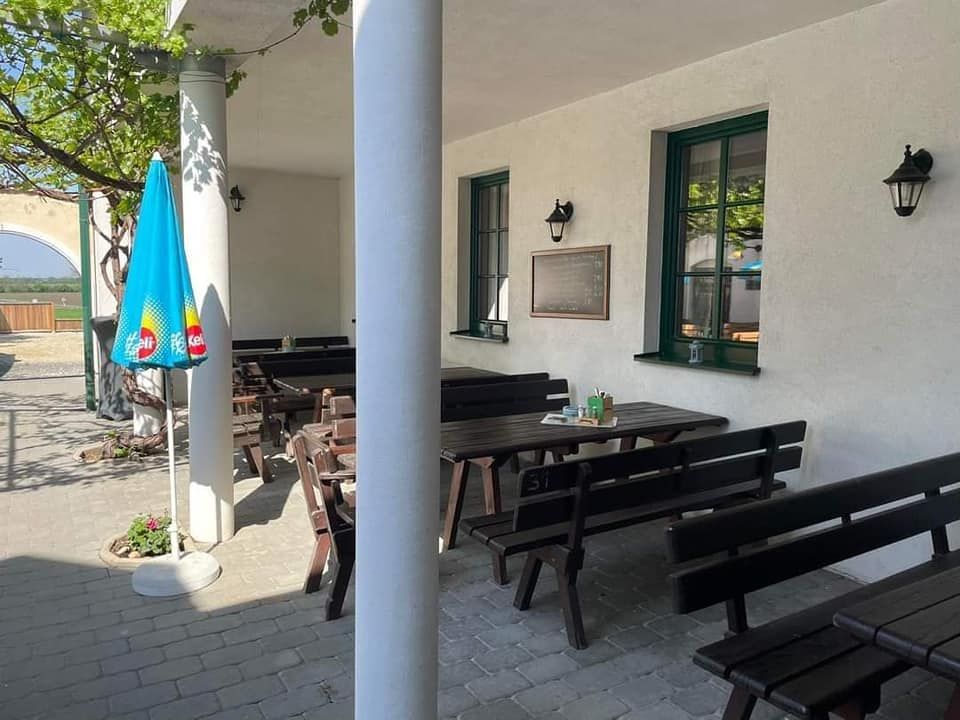 Egy üres vendégkert fapadokkal és asztalokkal, egy napernyővel és egy fehér ablakos fallal.