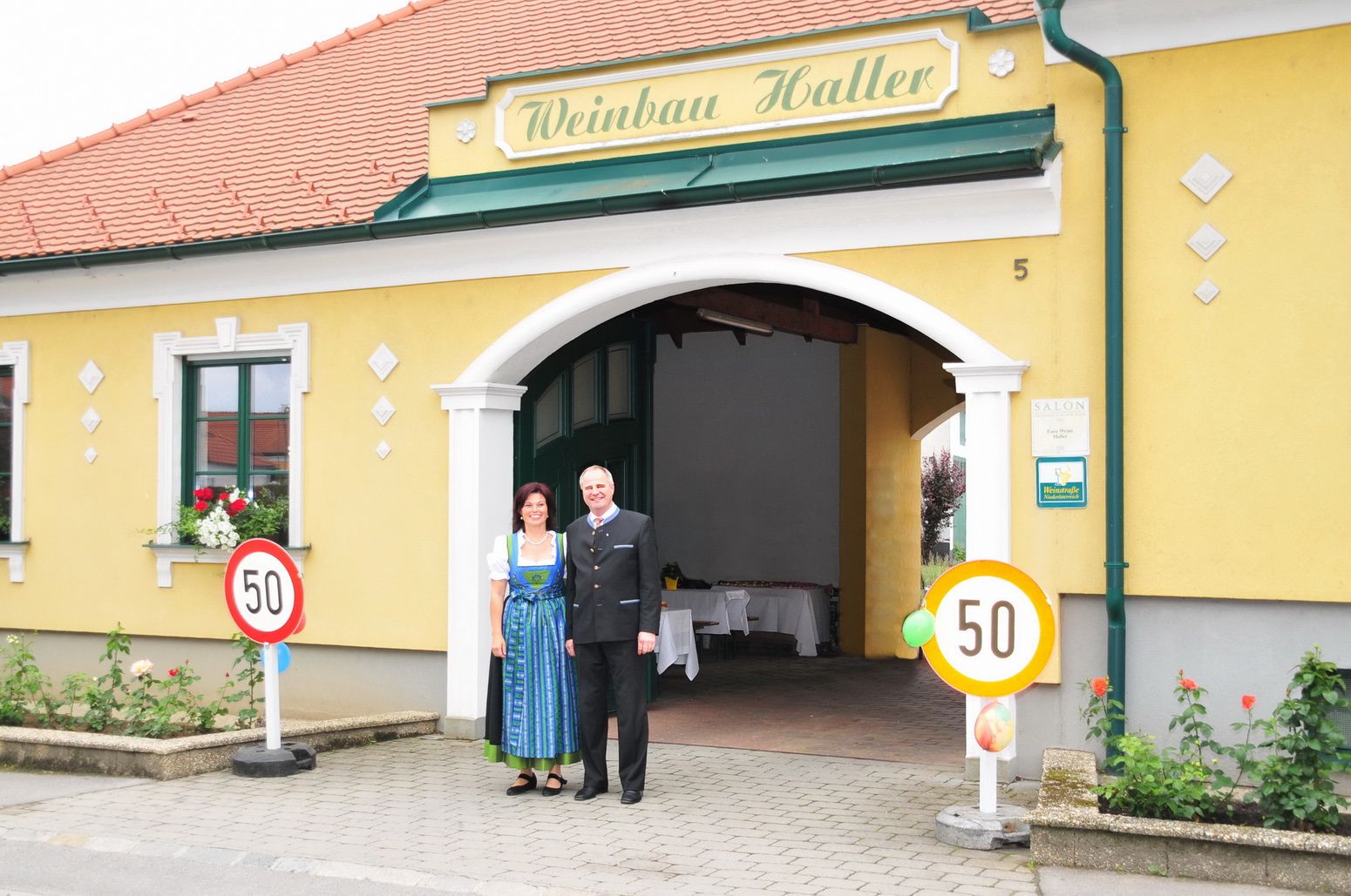 Egy pár áll egy sárga, "Weinbau Haller" feliratú épület előtt.
