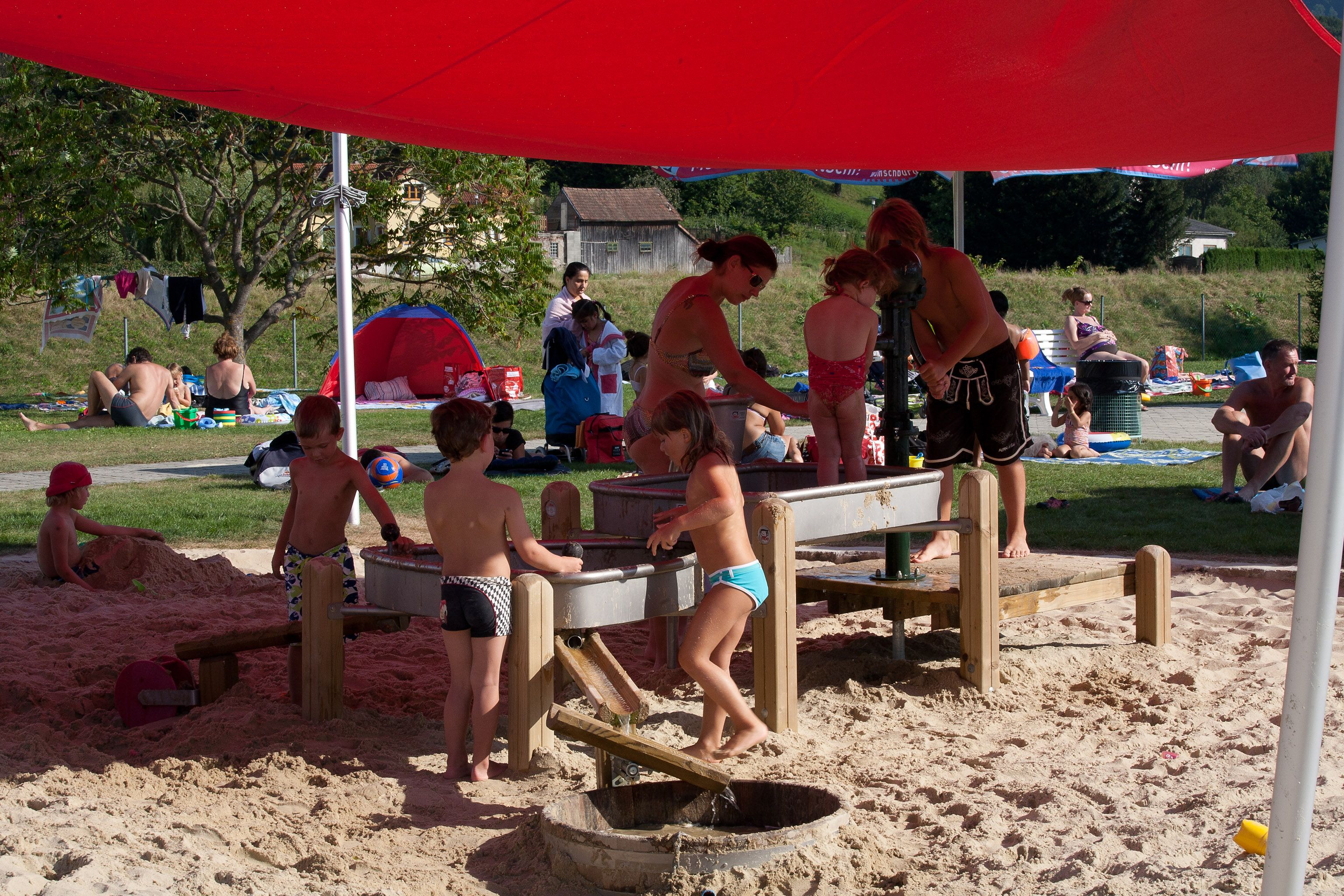 Gyerekek játszanak a homokban egy piros napellenző alatt a Wilhelmsburg Parkbadban.